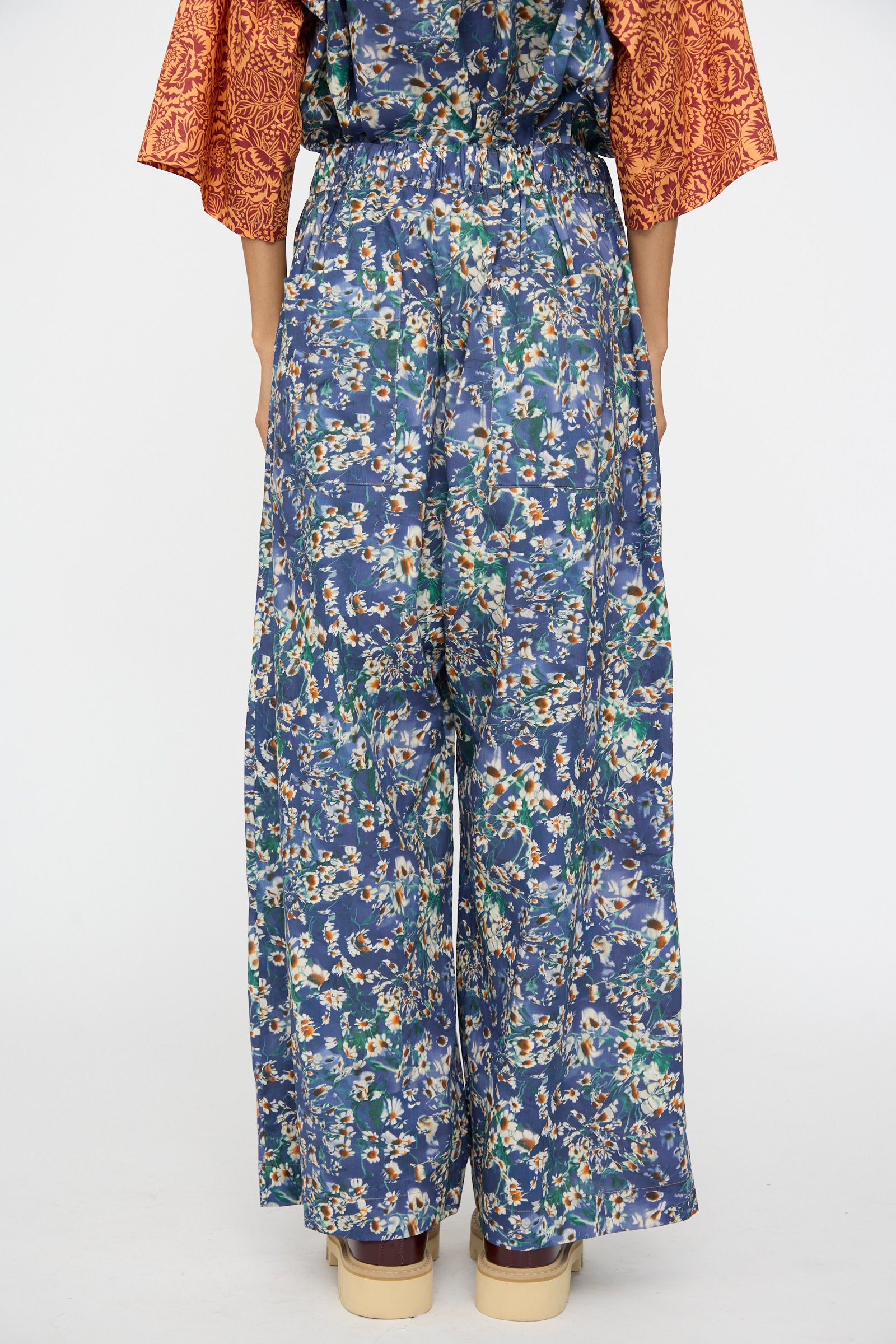 Person stands with back to camera, wearing KasMaria's Liberty Floral Baggy Pleated Trouser in Blue, paired with a top featuring orange textured sleeves—crafted from luxurious Liberty of London Tana Lawn Cotton.