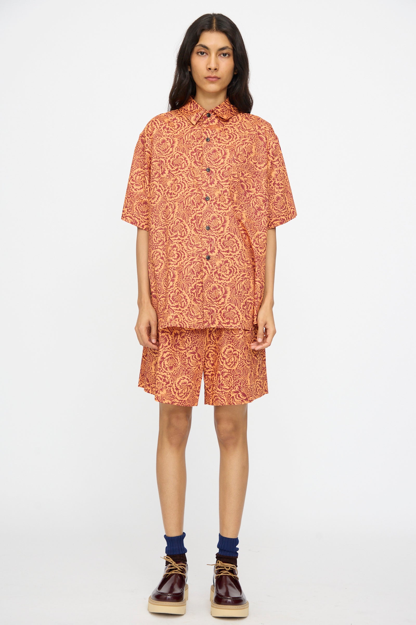 A person stands facing forward in the KasMaria Liberty Floral Classic Oversized Shirt in Orange, featuring an intricate pattern, paired with matching shorts, navy socks, and brown shoes against a plain white background.