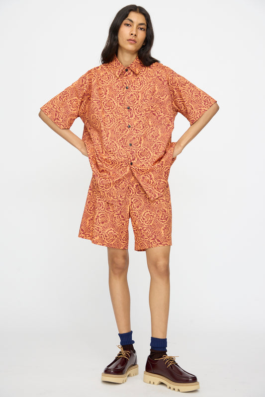 A person stands with hands on hips, wearing the KasMaria Liberty Floral Classic Oversized Shirt in Orange and matching shorts, paired with blue socks and brown platform shoes.