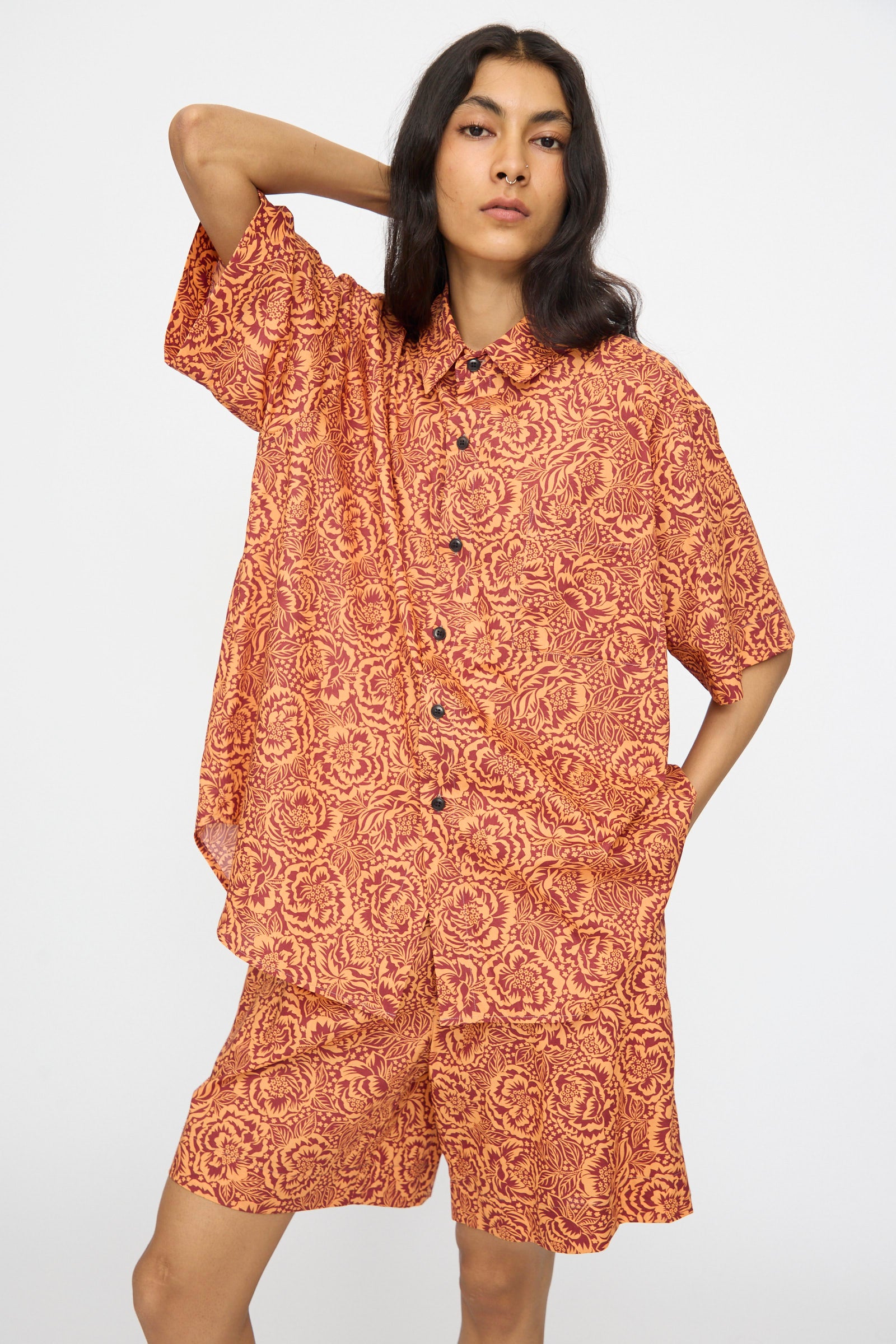 A person poses against a plain background wearing the KasMaria Liberty Floral Classic Oversized Shirt in Orange, featuring an oversized fit and bold orange and red floral pattern crafted from Liberty of London’s Tana Lawn Cotton.