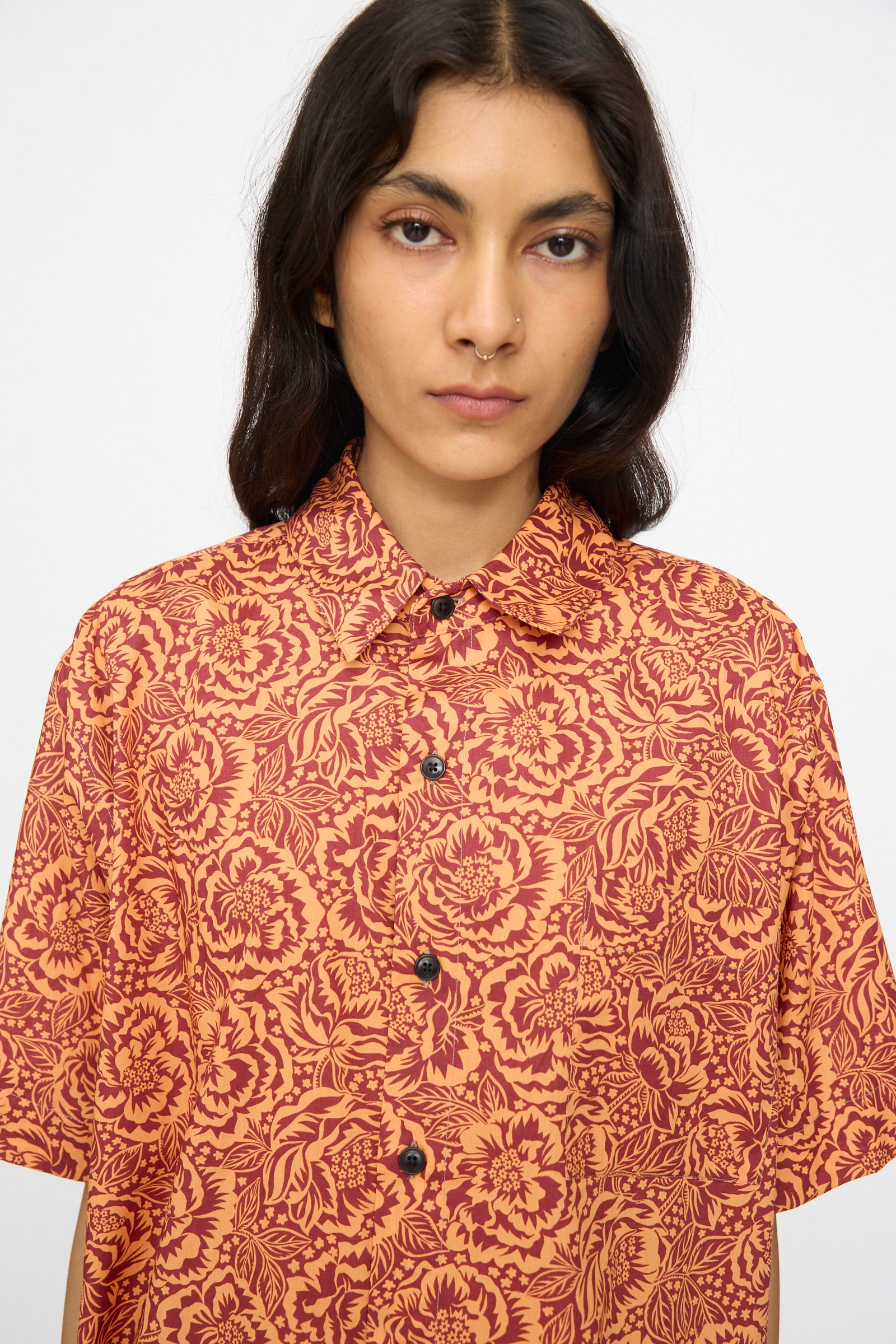 A person with long dark hair wears the KasMaria Liberty Floral Classic Oversized Shirt in Orange, crafted from Liberty of London Tana Lawn Cotton, while standing against a plain light background.