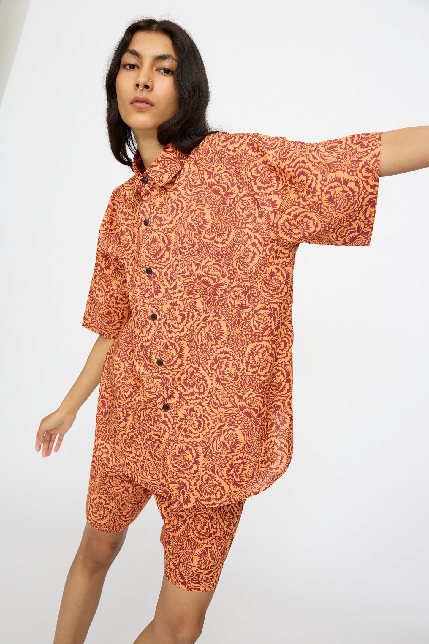 Someone stands against a plain white background wearing the KasMaria Liberty Floral Classic Oversized Shirt in Orange, featuring an oversized fit and a red and orange floral pattern crafted from Liberty of London Tana Lawn Cotton.