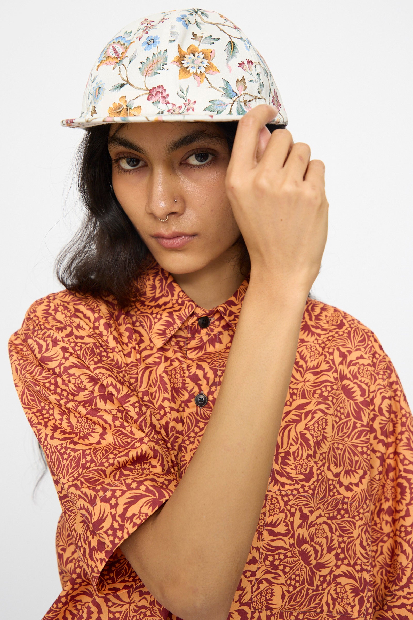 A person faces the camera, one hand adjusting their KasMaria Liberty Floral Long Bill Cap, pairing it with an orange patterned shirt.