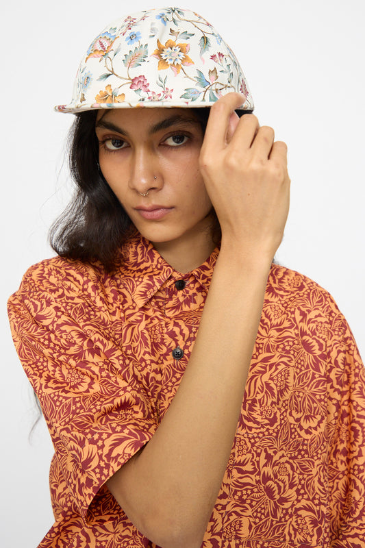A person faces the camera, one hand adjusting their KasMaria Liberty Floral Long Bill Cap, pairing it with an orange patterned shirt.
