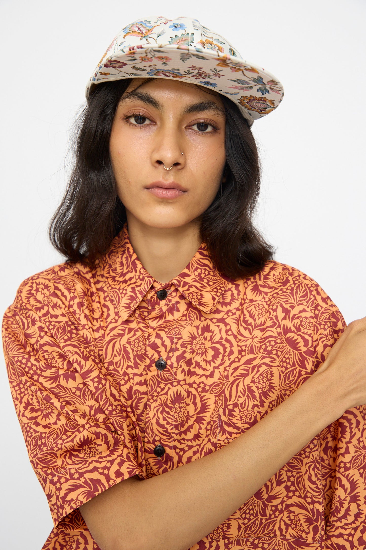A person wears a patterned orange shirt and the KasMaria Liberty Floral Long Bill Cap, crafted from a soft cotton linen blend, looking at the camera with a neutral expression against a plain background.