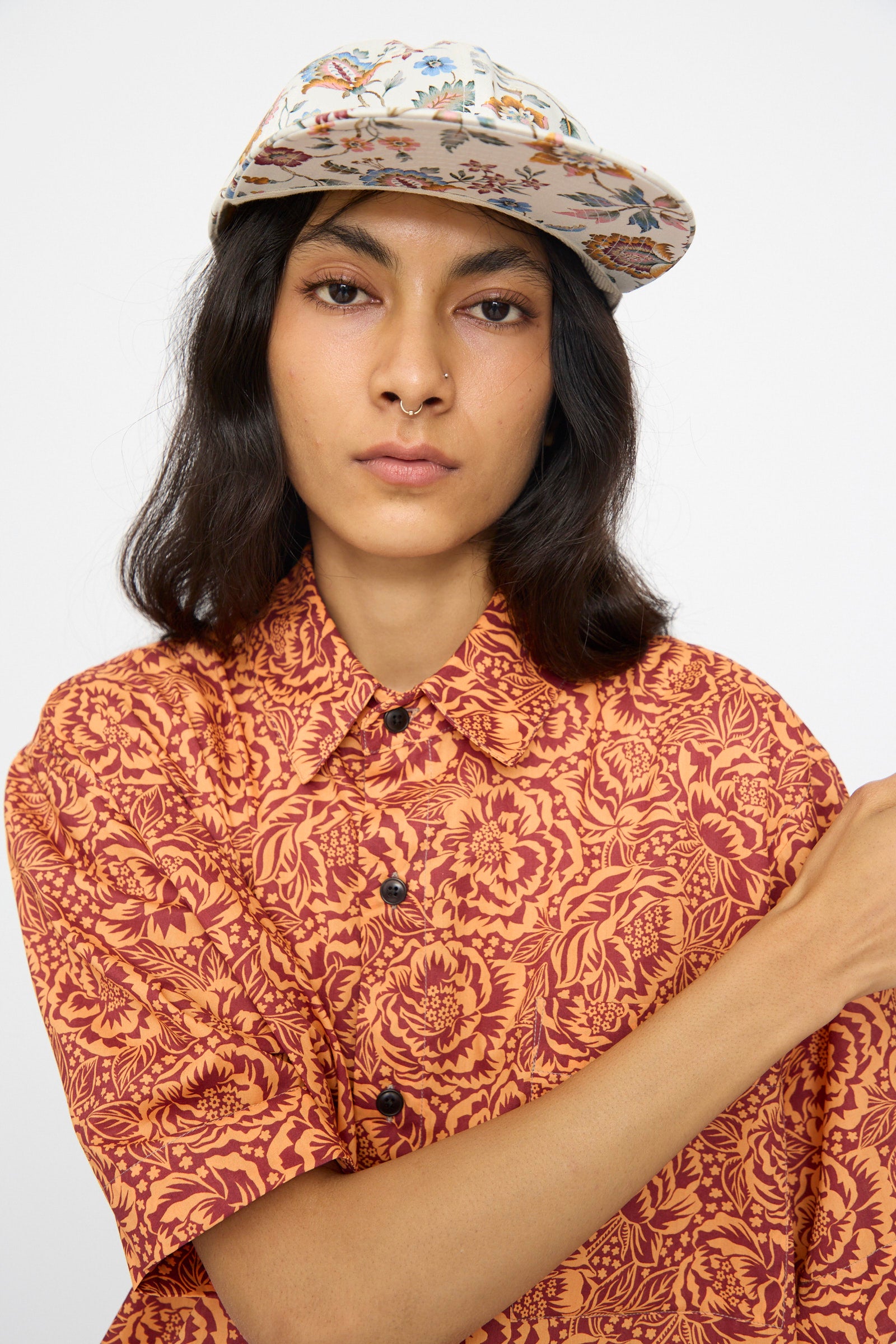 A person wears a patterned orange shirt and the KasMaria Liberty Floral Long Bill Cap, crafted from a soft cotton linen blend, looking at the camera with a neutral expression against a plain background.