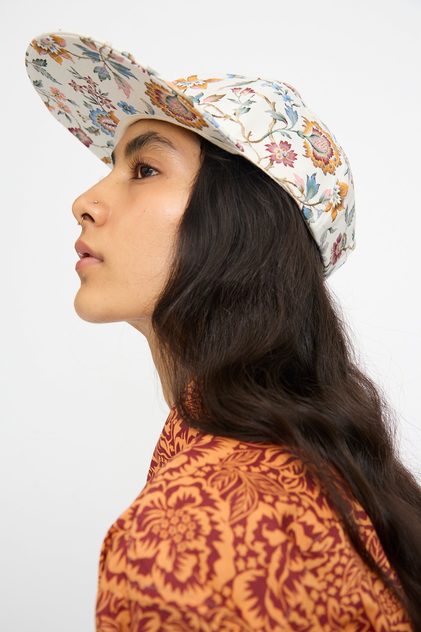 A person with long dark hair poses against a plain background wearing a cotton-linen orange floral shirt and the KasMaria Liberty Floral Long Bill Cap.