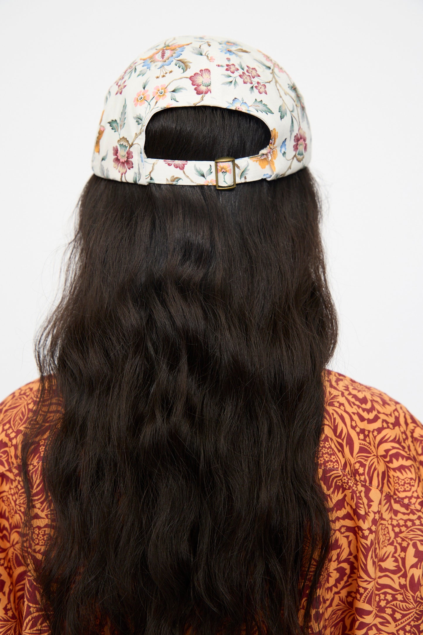 A person with long, wavy dark hair, seen from behind, wears the KasMaria Liberty Floral Long Bill Cap and an orange garment featuring a red decorative design.