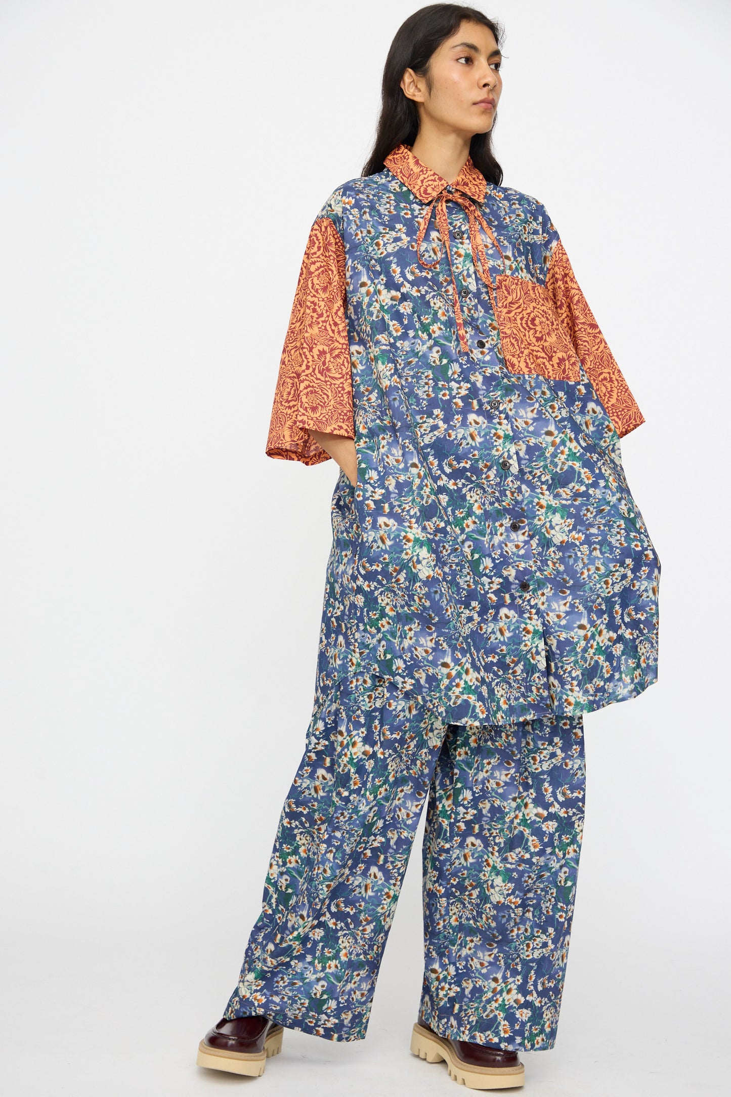 A person wears the KasMaria Liberty Floral Oversized Shirtdress in Blue and Orange, featuring blue florals with contrasting red-orange accents, standing against a plain background.