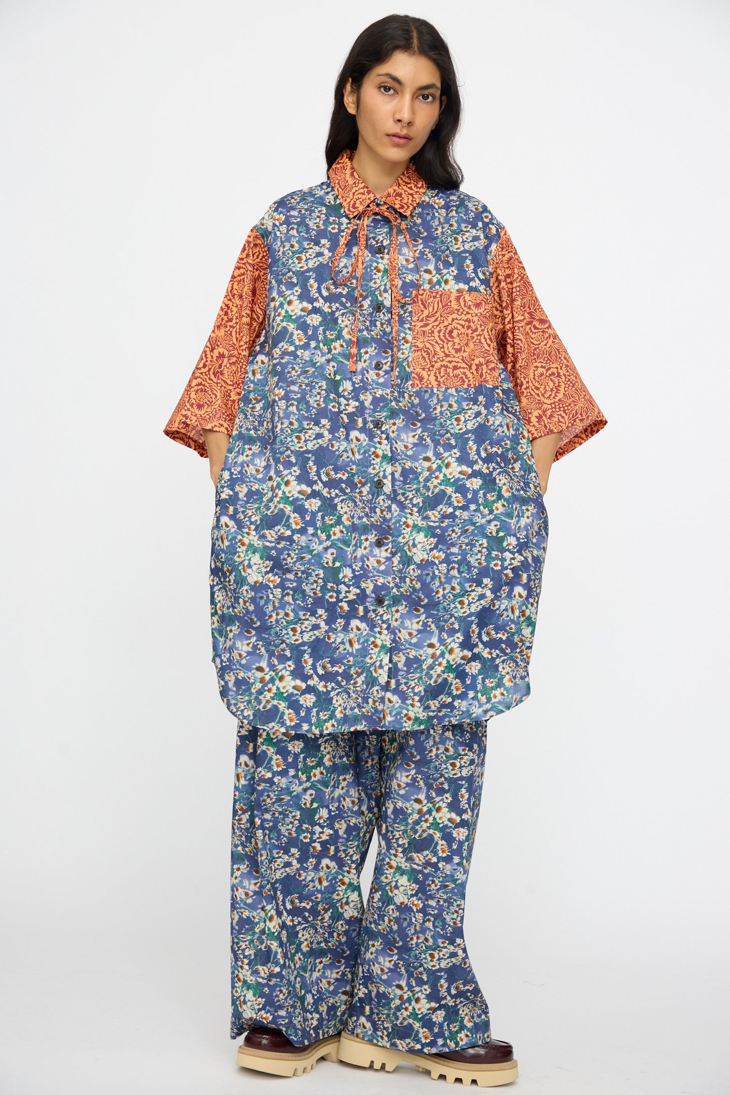 A person wears the KasMaria Liberty Floral Oversized Shirtdress in Blue and Orange, featuring mixed patterns, paired with beige platform shoes.