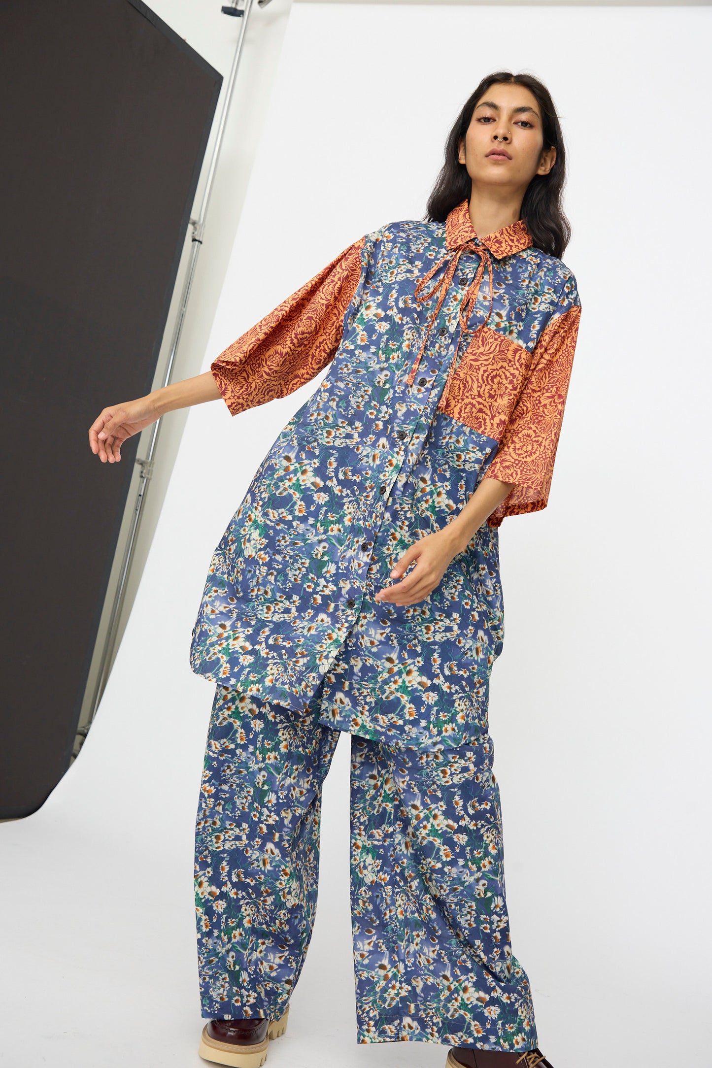 A person poses confidently in a studio wearing the KasMaria Liberty Floral Oversized Shirtdress in Blue and Orange, inspired by Liberty of London prints, against a white backdrop.