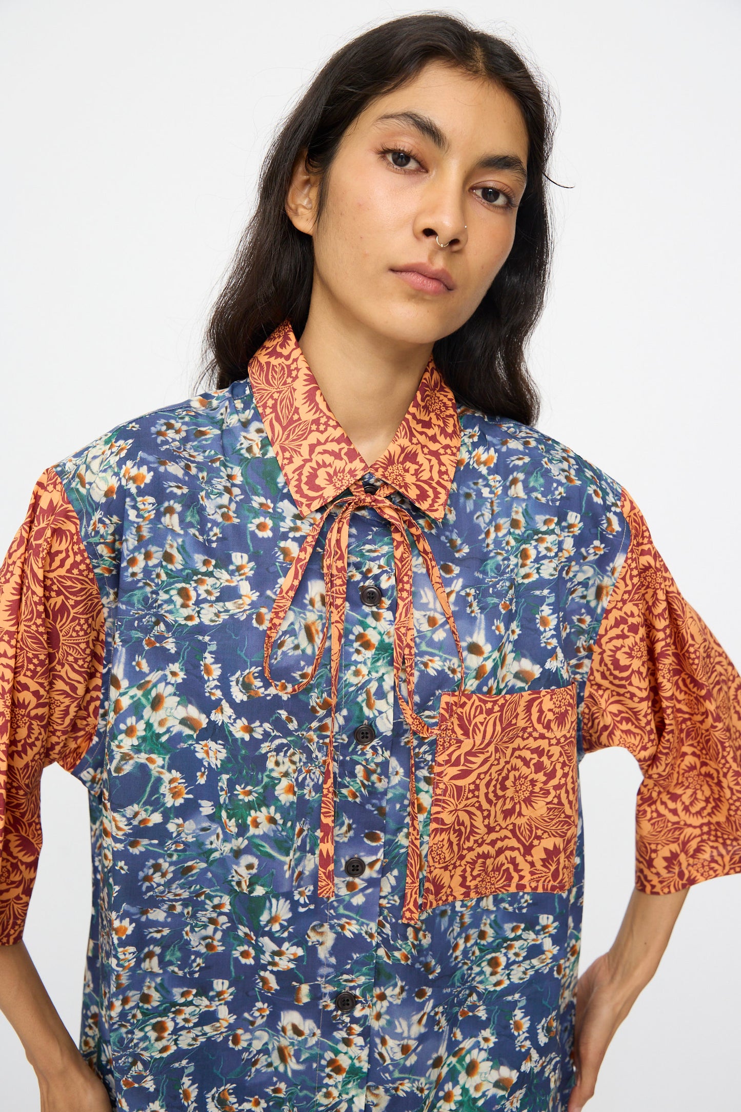 A woman wears the KasMaria Liberty Floral Oversized Shirtdress in Blue and Orange, featuring a blue floral pattern with contrasting orange collar, sleeves, and pocket, standing against a plain background.