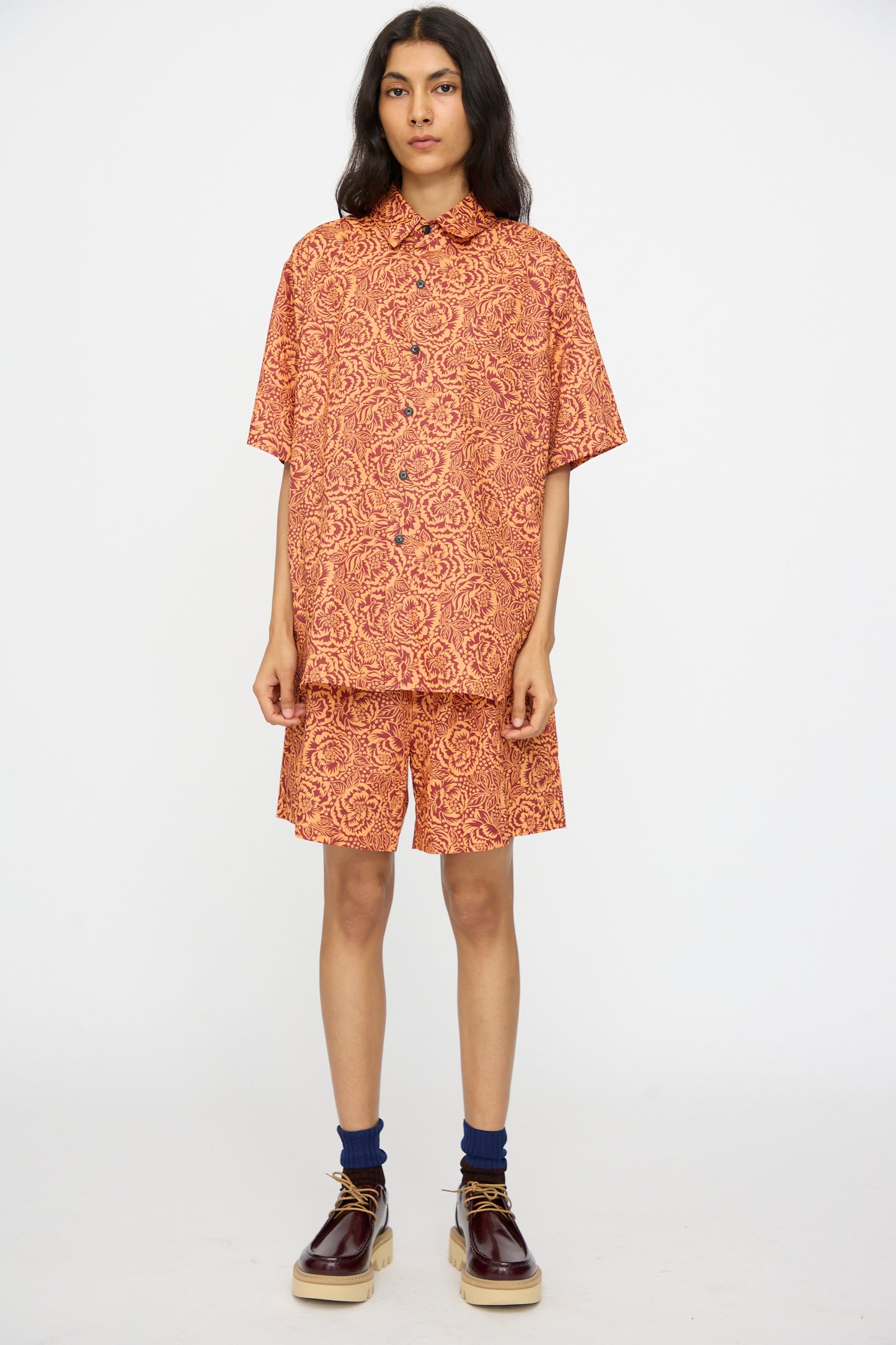 A person stands against a plain background wearing KasMaria's Liberty Floral Shorts in Orange by Liberty of London, paired with a matching patterned short-sleeve shirt, navy socks, and brown platform shoes.