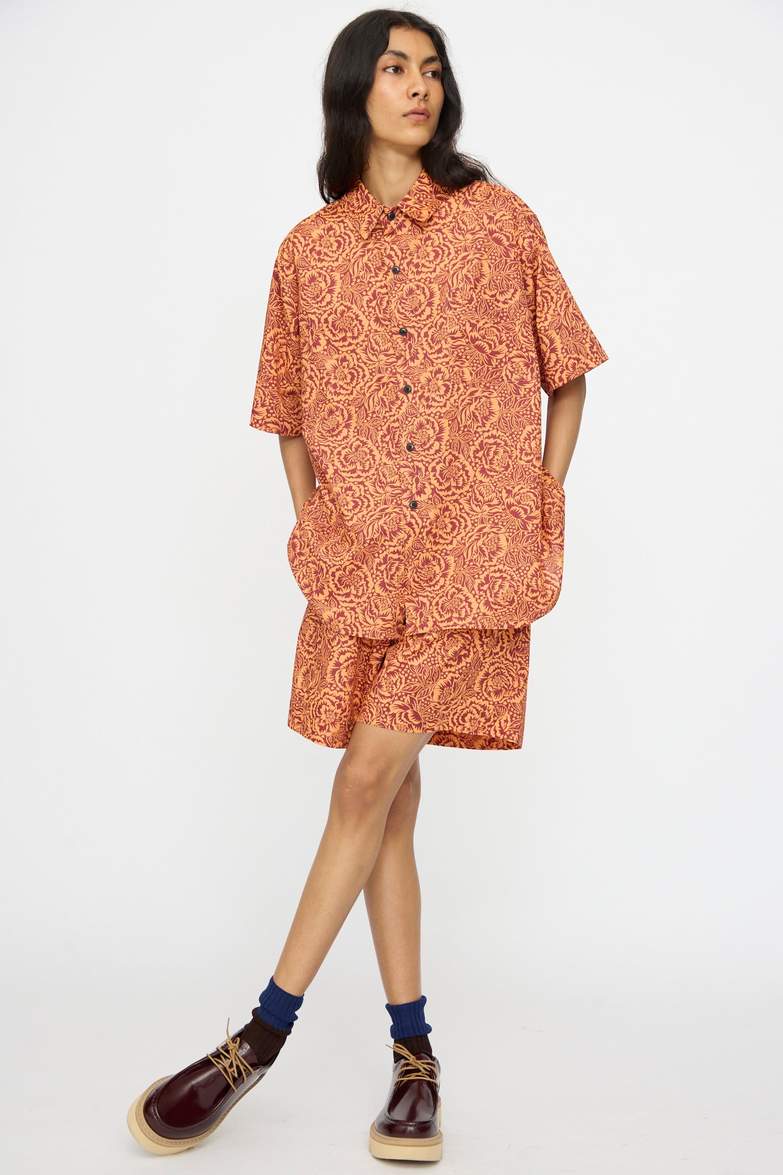 A person stands against a plain white background wearing KasMaria Liberty Floral Shorts in Orange with a matching short-sleeve shirt, navy socks, and burgundy shoes.