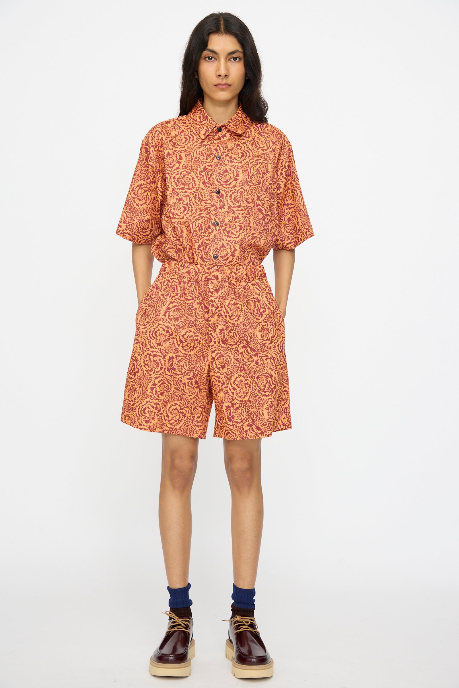 A person stands against a plain background wearing KasMaria Liberty Floral Shorts in Orange made from Liberty of London Tana Lawn Cotton, paired with a matching short-sleeve shirt, blue socks, and dark brown shoes.