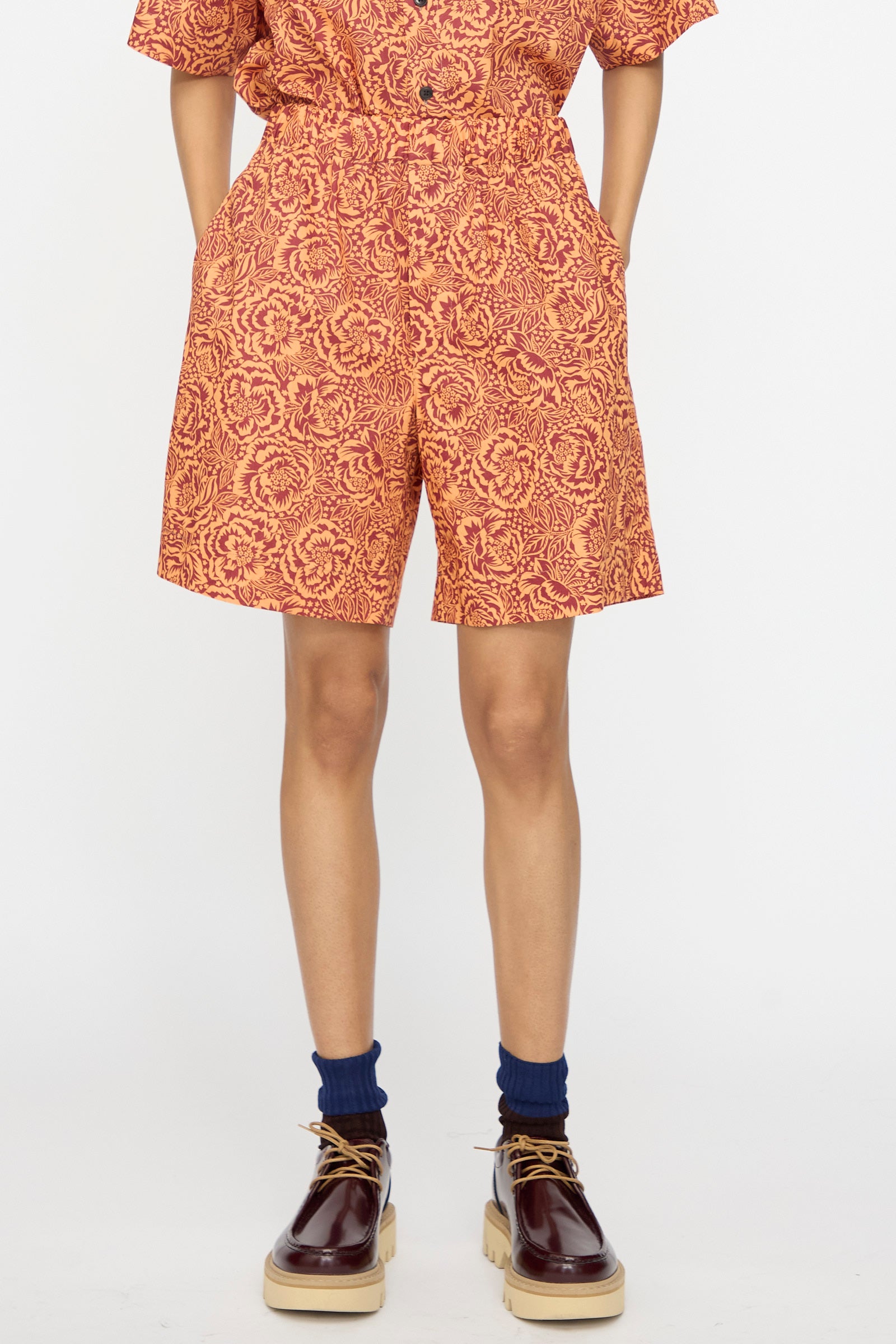 A person wears KasMaria Liberty Floral Shorts in Orange with a matching top, navy socks, and brown platform shoes, standing against a plain white background.