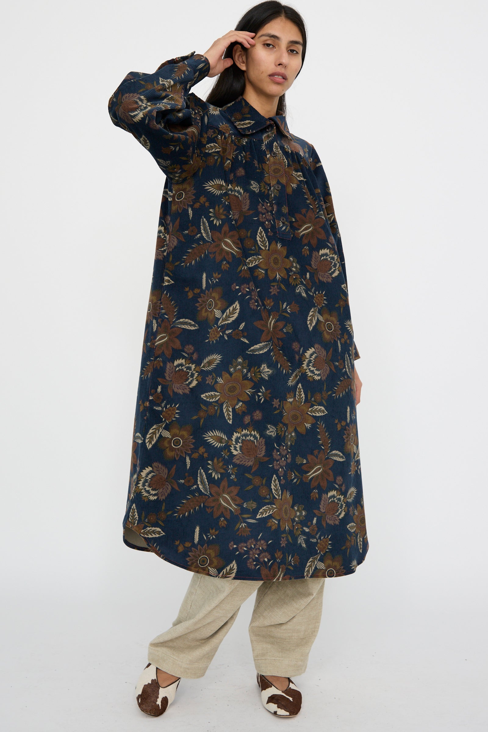 A person wearing the KasMaria Corduroy Dress in Printed Floral design over beige pants, paired with white and brown shoes, is standing against a plain background with their arm raised
.