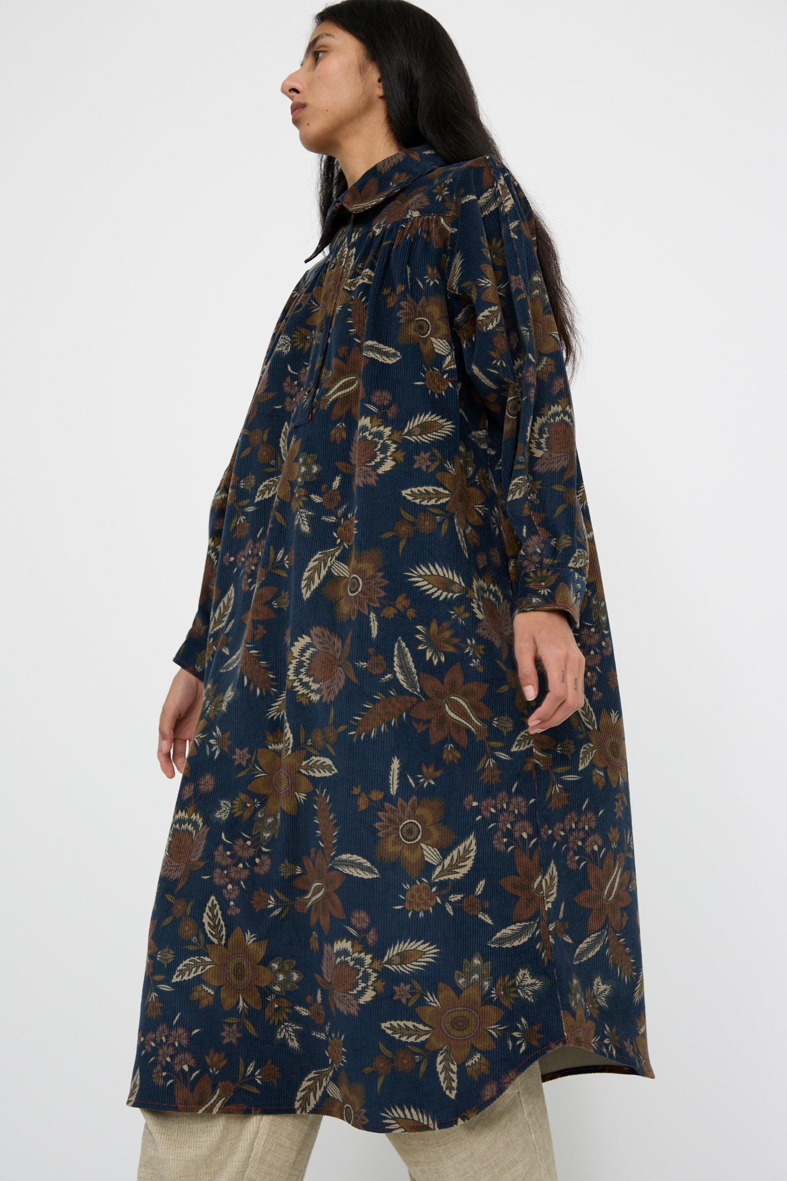 A person wears the KasMaria Corduroy Dress in Printed Floral, a long dark blue shirt dress, paired with beige pants while standing against a plain background, walking to the left.