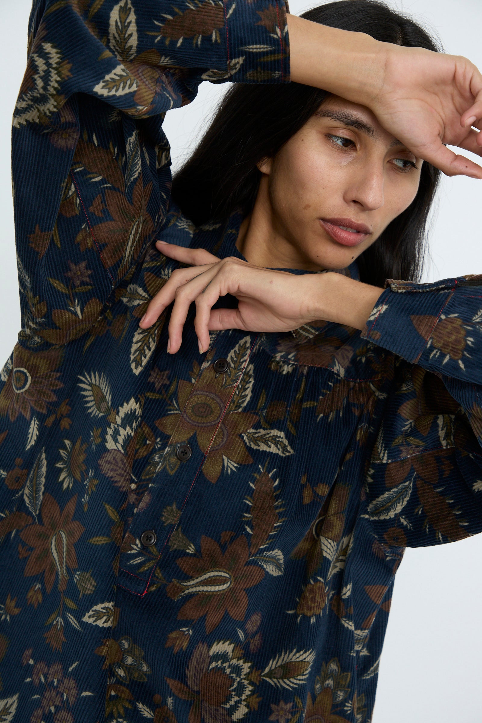 A person wearing the Corduroy Dress in Printed Floral by KasMaria, crafted from 100% cotton, poses with one arm raised and the other across their chest.