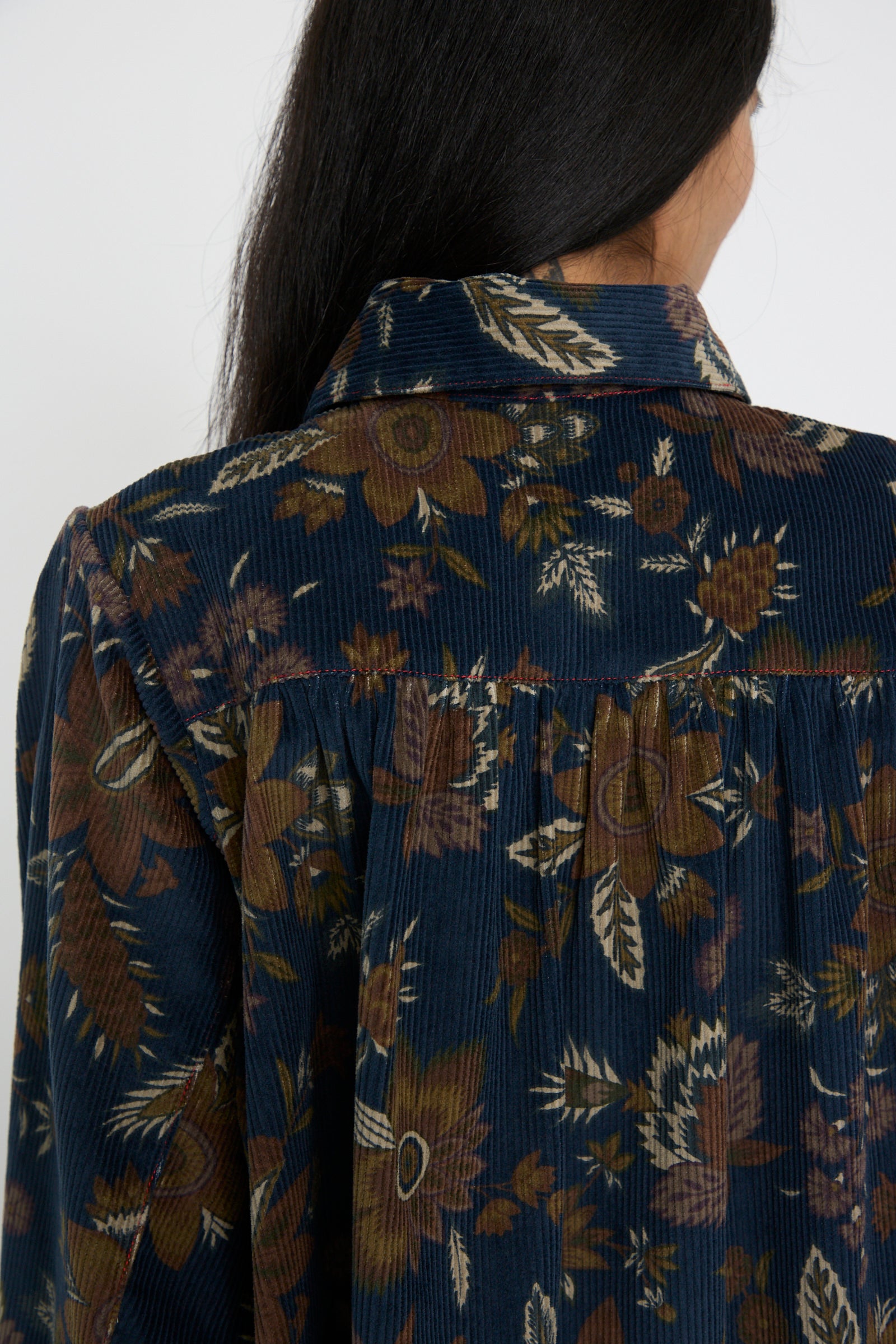 Rear view of a person with long dark hair wearing the Corduroy Dress in Printed Floral by KasMaria, crafted from 100% cotton and featuring a delicate floral pattern in brown and beige tones.