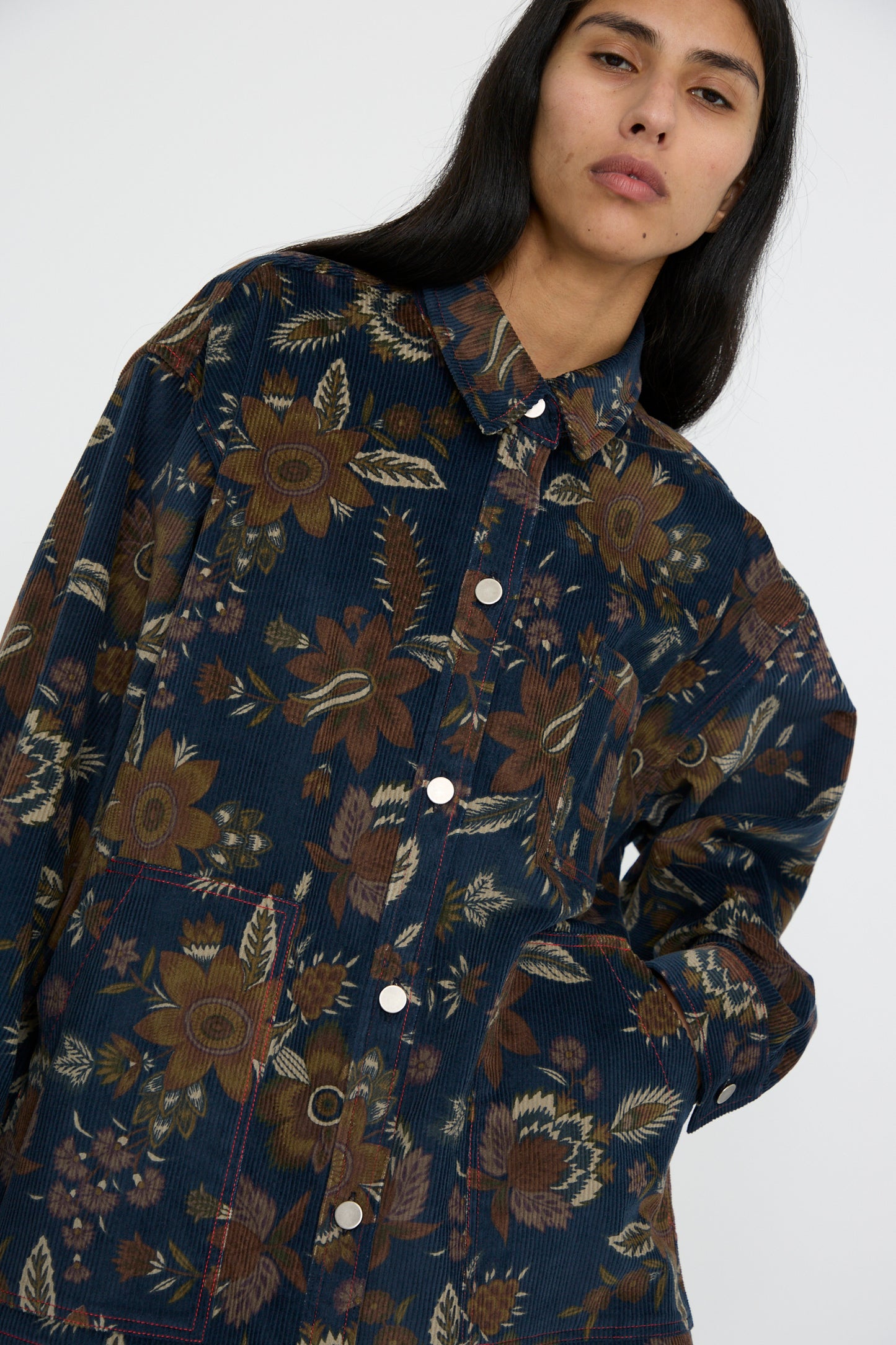 A person wearing the KasMaria Corduroy Overshirt in Printed Floral stands against a plain background, facing forward with their hand in a pocket.