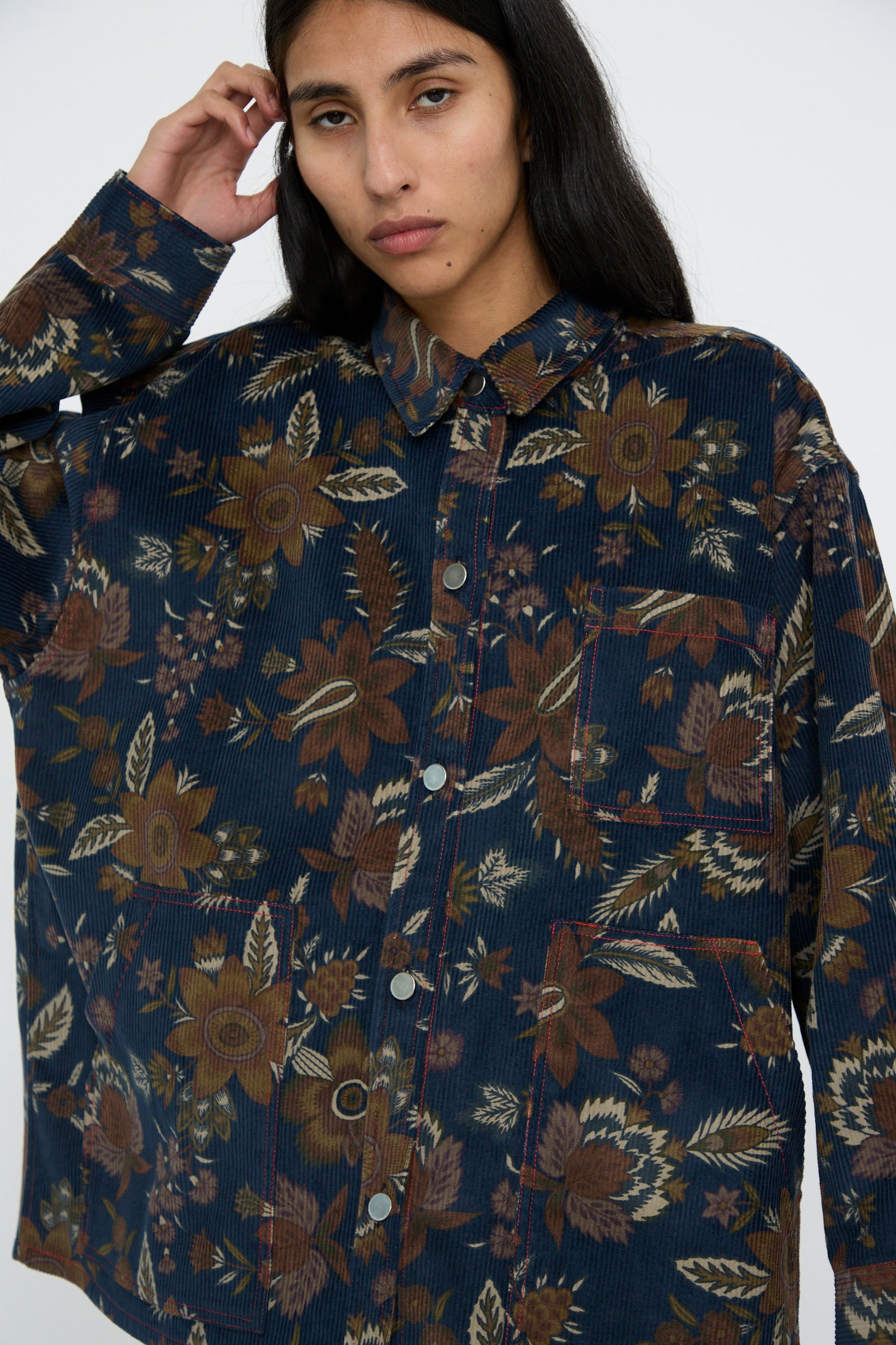 A person wearing the KasMaria Corduroy Overshirt in Printed Floral stands against a plain background with one hand touching their hair.