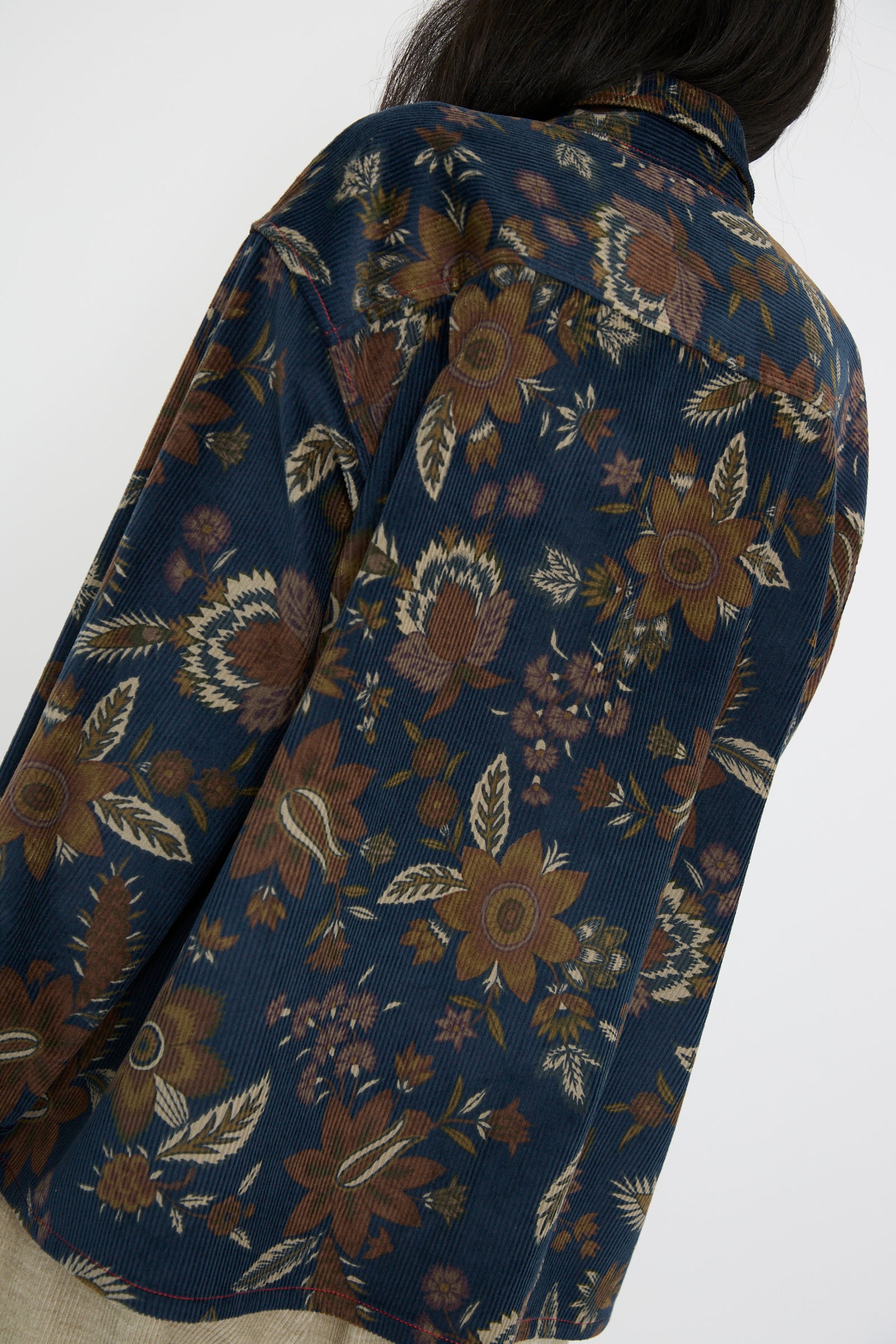 A person seen from the back is wearing the KasMaria Corduroy Overshirt in Printed Floral, featuring a Japanese floral pattern in earthy tones, against a white background. 