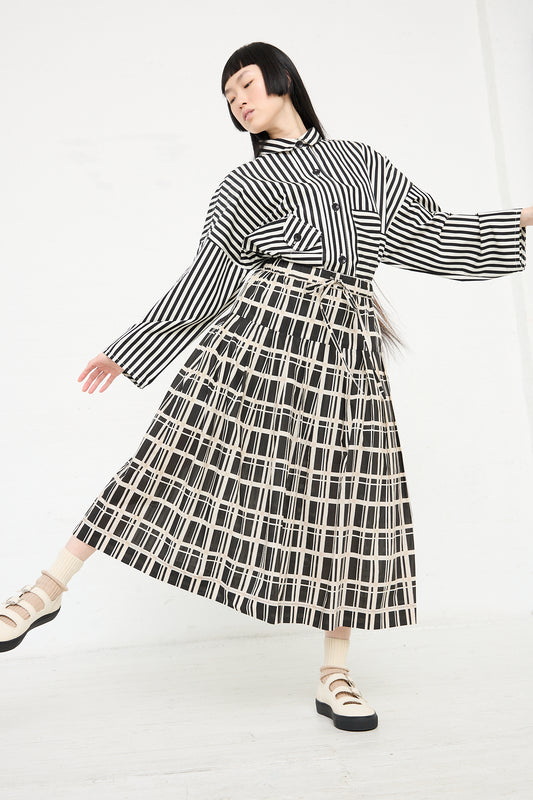 A woman posing in a KasMaria Cotton Pleated Long Skirt in Grid Print and a black and white striped, relaxed fit shirt, with one arm extended, against a white backdrop.