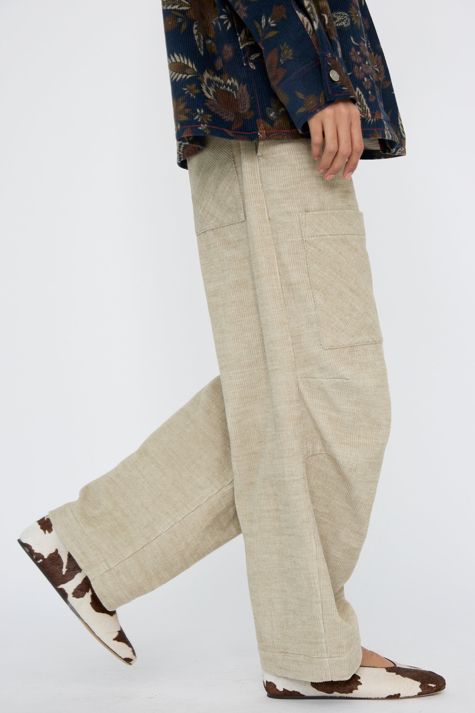 A person wearing KasMaria's Cotton Wool Corduroy Pant with Front Patch in beige, paired with a floral patterned shirt and cow-print shoes, walks gracefully against a plain background.