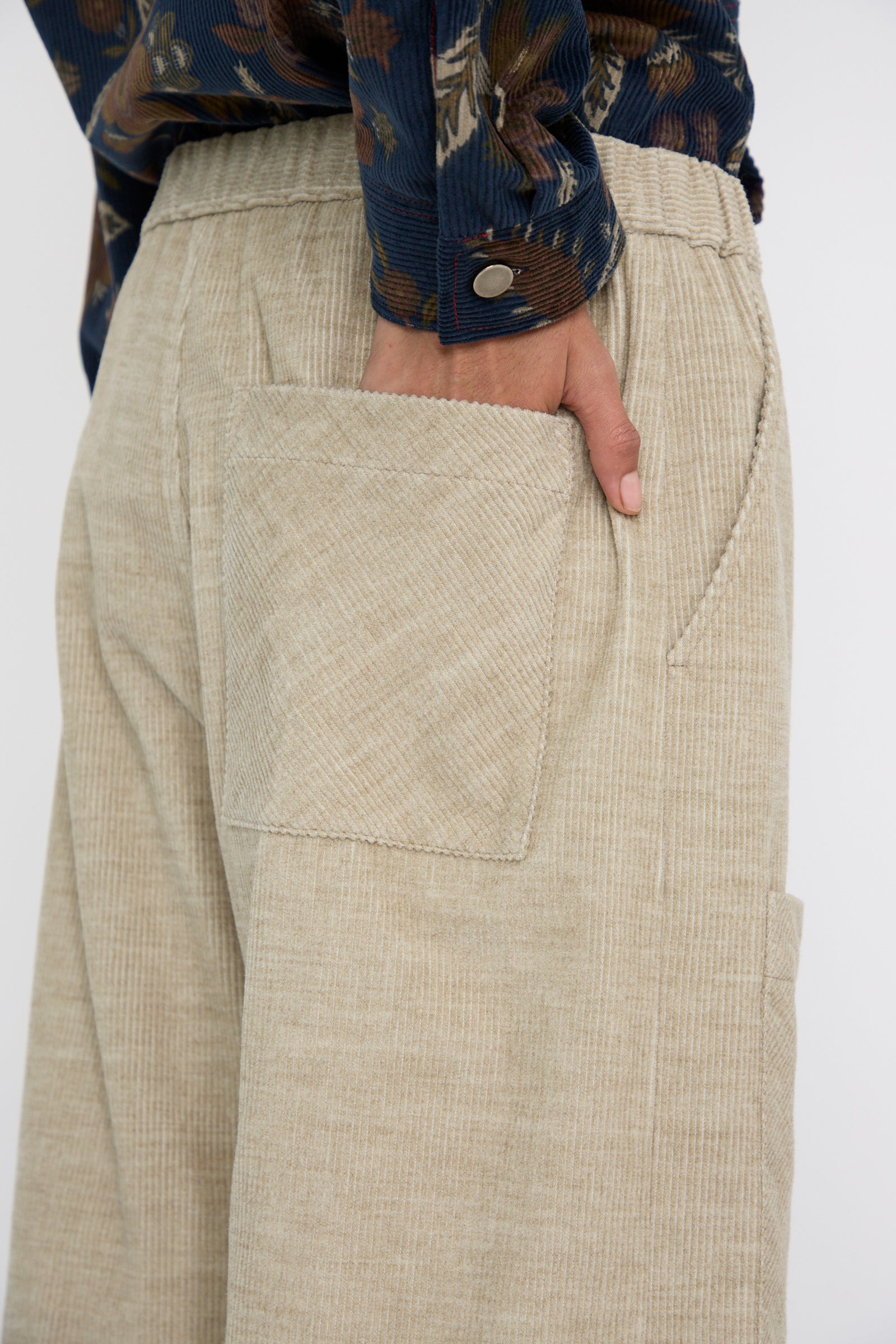 A close up of a person wearing KasMaria's Cotton Wool Corduroy Pant with Front Patch in beige, featuring an elasticated waist and a hand in the back pocket, paired with a patterned shirt.