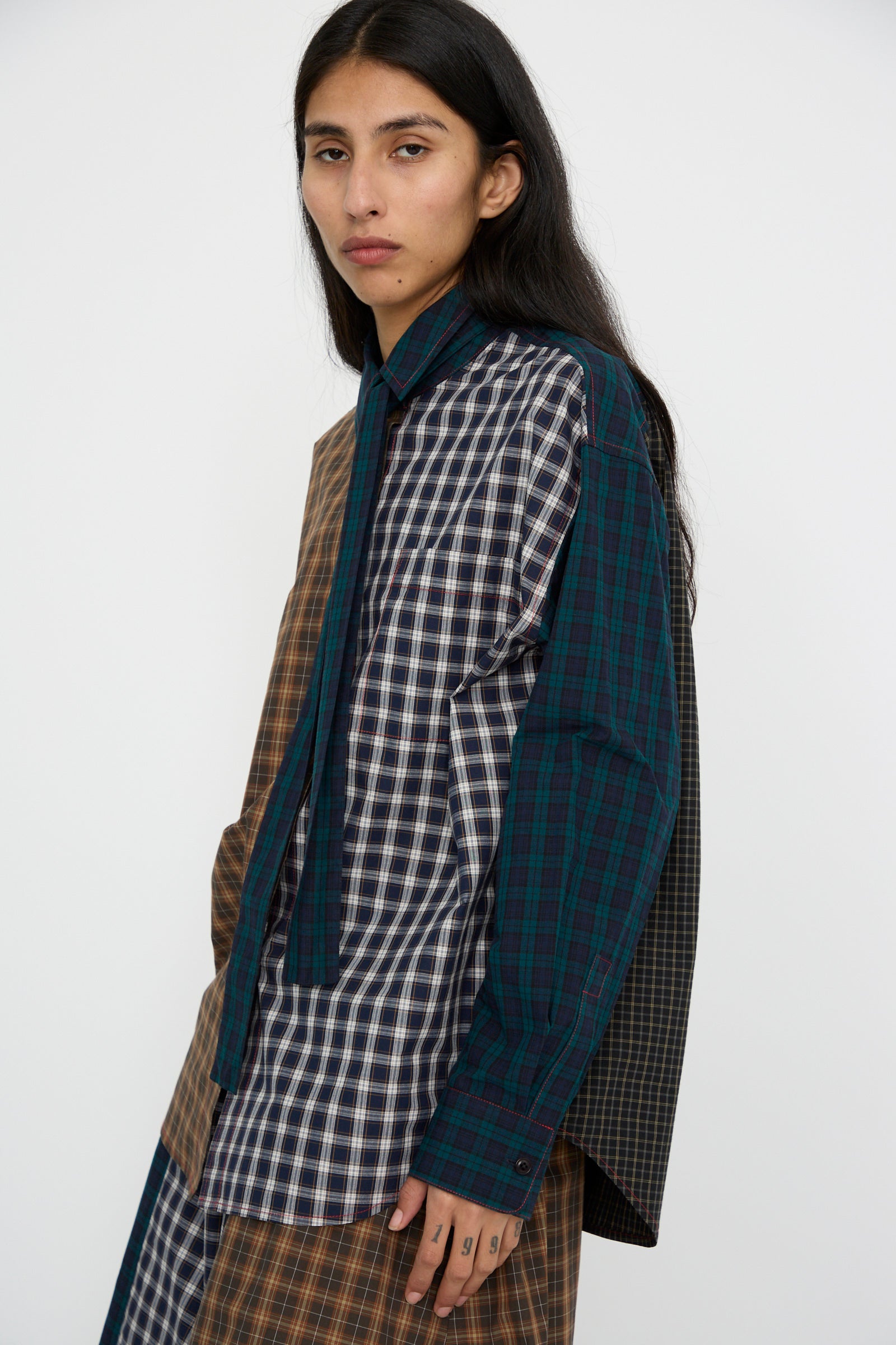 A person with long dark hair is wearing the KasMaria Heavy Poplin Oversized Shirt with Tie in Mixed Plaid, standing against a plain background.