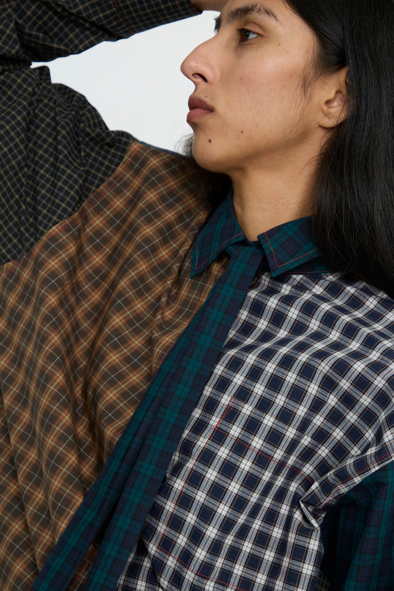 A close up of a person looking in profile and wearing the Heavy Poplin Oversized Shirt with Tie in Mixed Plaid by KasMaria, featuring a combination of brown, blue, and green plaids and enhanced with Italian Corozo buttons.