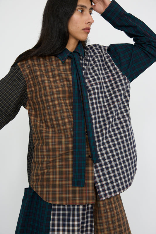 A person is wearing the Heavy Poplin Oversized Shirt with Tie in Mixed Plaid by KasMaria, featuring a long-sleeve design in brown, green, and navy crafted from Japanese cotton poplin, looking to the left.