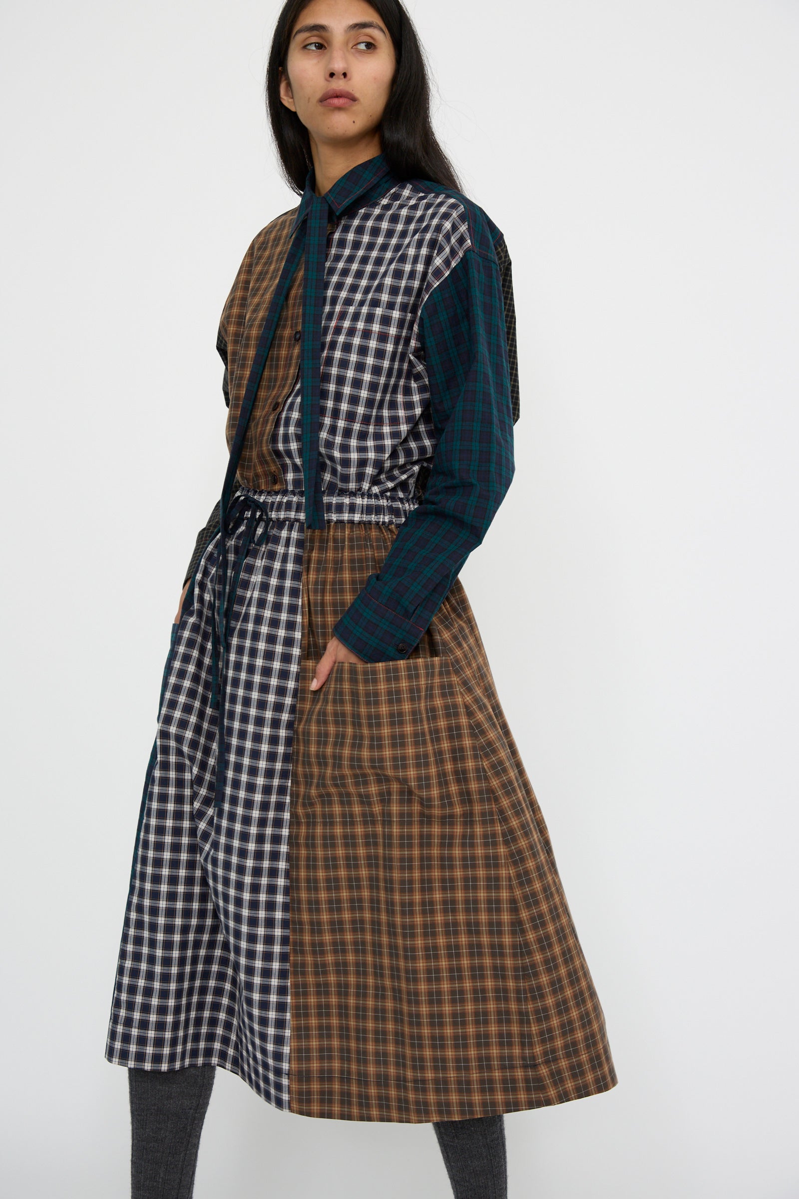 A person wearing the KasMaria Heavy Poplin Skirt in Mixed Plaid, featuring a brown and blue plaid design made from Japanese cotton, stands against a plain white background.