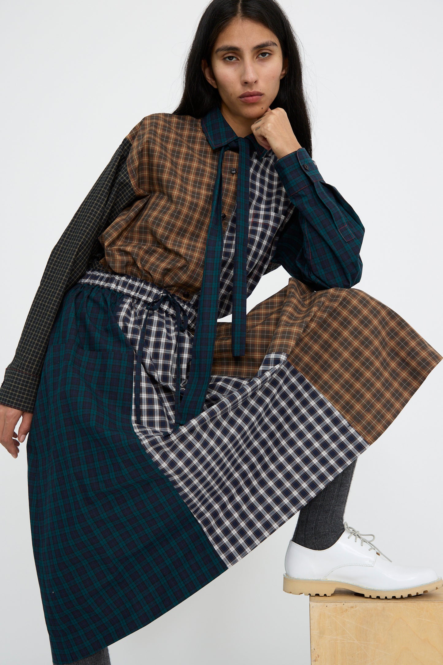 A person styled in the KasMaria Heavy Poplin Skirt in Mixed Plaid, made from Japanese cotton, paired with white shoes, poses with one knee on a wooden block against a plain backdrop.