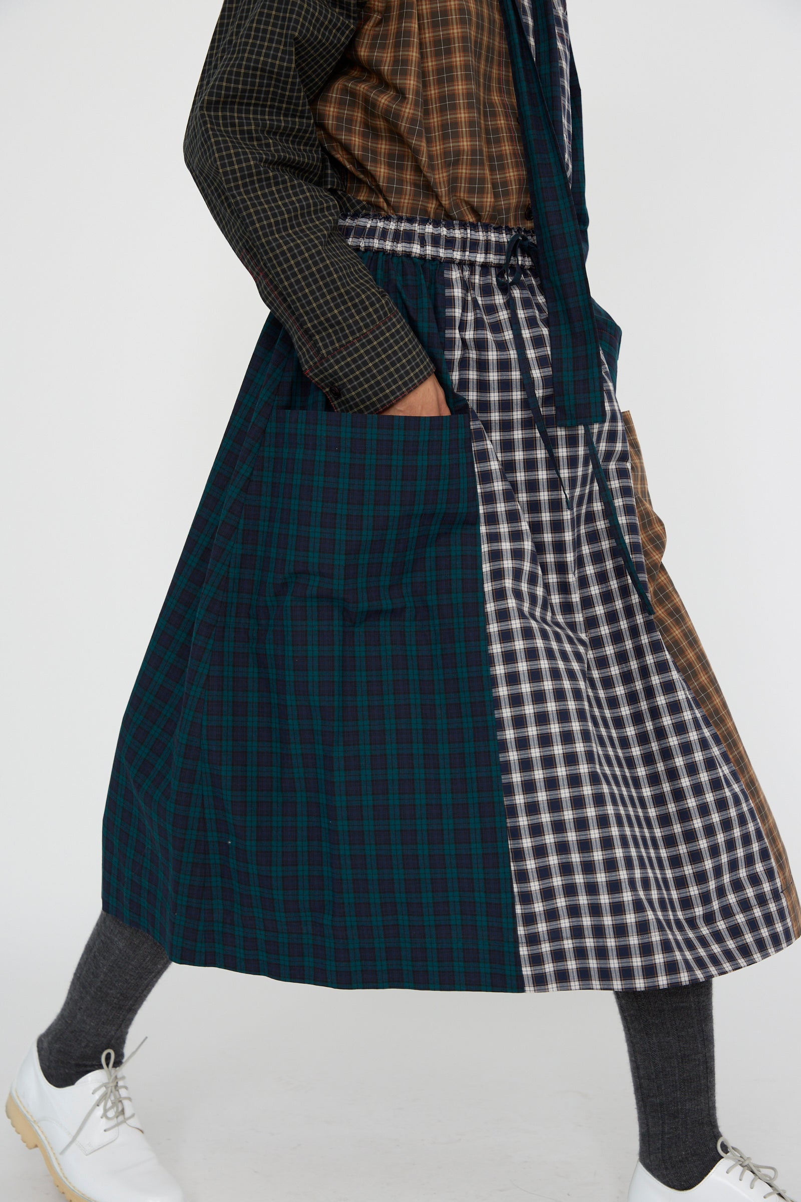 A person dressed in KasMaria's Heavy Poplin Skirt in Mixed Plaid, showcasing a classic plaid design, with hands tucked into the pockets, paired with gray socks and white shoes against a plain background.