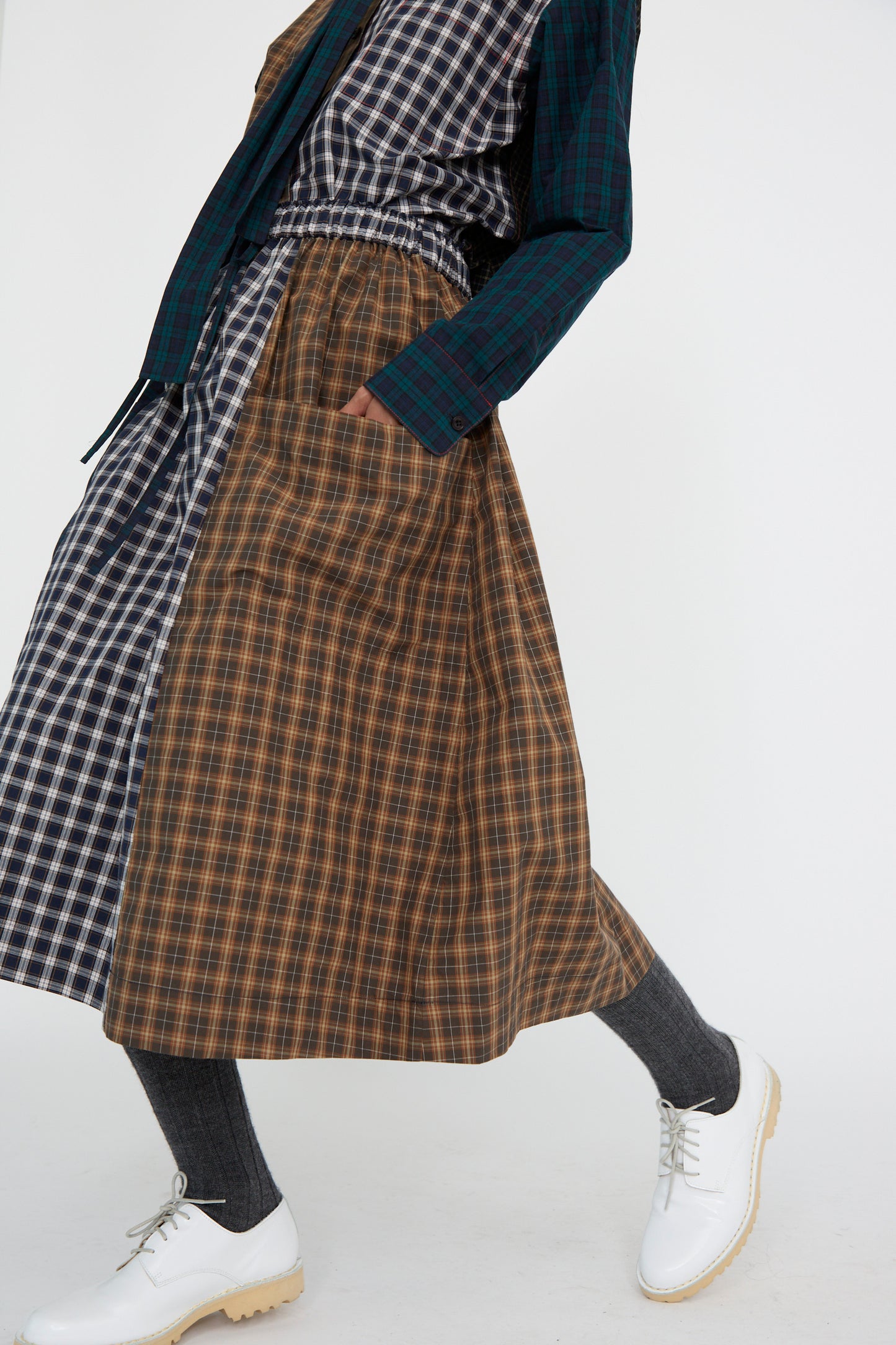 A person dressed in KasMaria's Heavy Poplin Skirt in Mixed Plaid, featuring a checkered pattern and paired with a long-sleeved Japanese cotton shirt, gray socks, and white shoes.