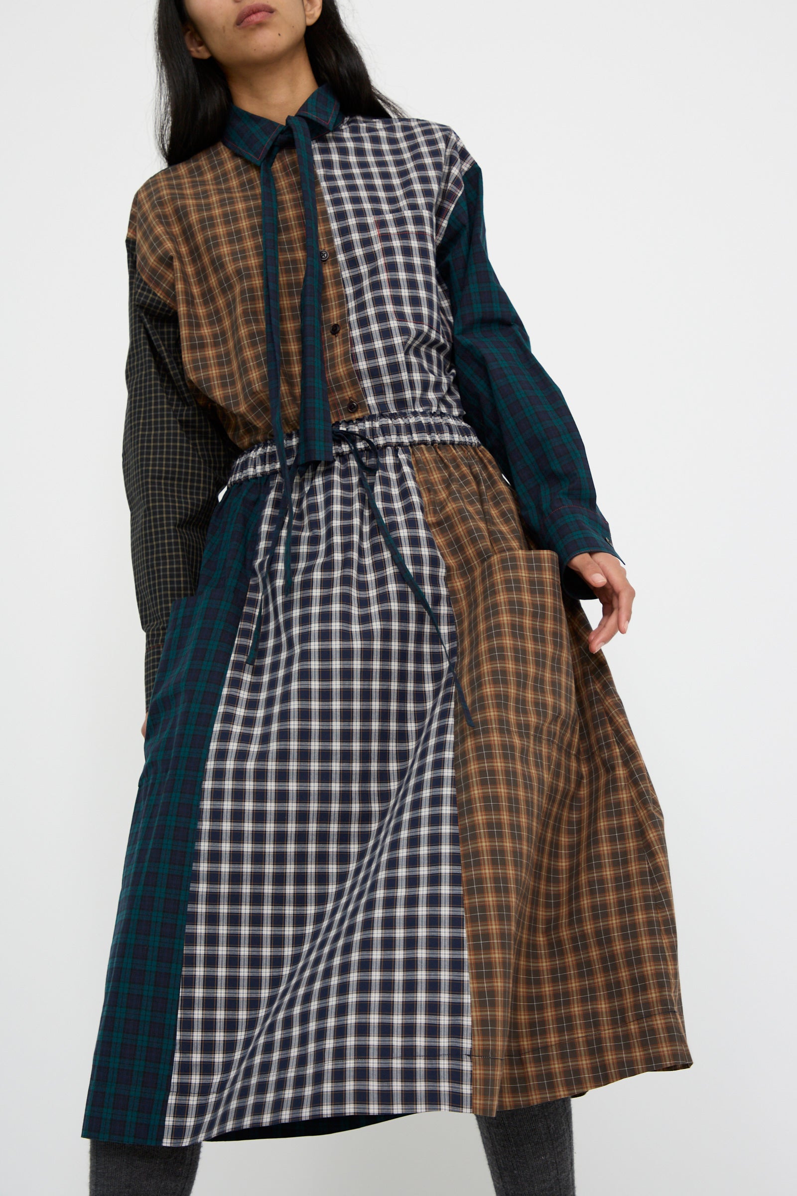 A person wearing a long-sleeve dress made from Japanese cotton with a mix of plaid patterns, paired with the Heavy Poplin Skirt in Mixed Plaid by KasMaria, stands against a plain white background.