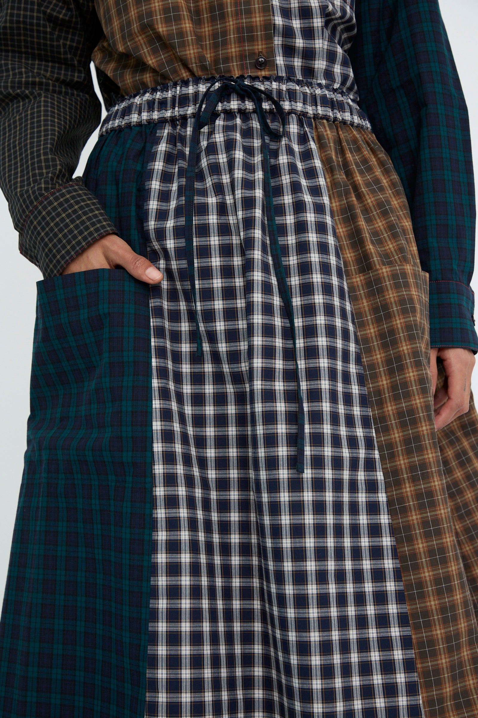 A close up of a person wearing the Heavy Poplin Skirt in Mixed Plaid from KasMaria, made from Japanese cotton with various patterns and colors, accessorized with a drawstring waist and a large side pocket. 