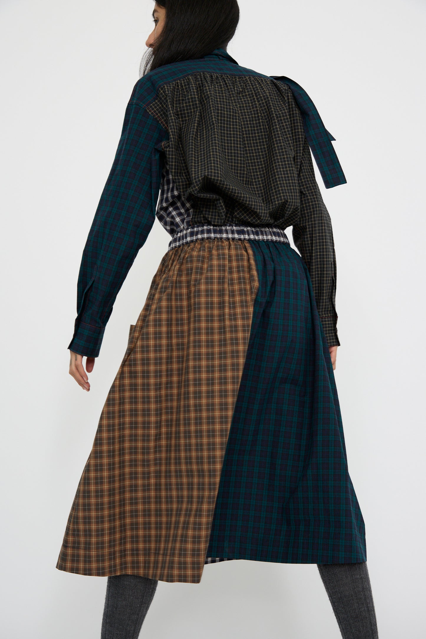 A person shown from the back stands against a plain white backdrop, wearing the Heavy Poplin Skirt in Mixed Plaid by KasMaria, featuring long sleeves and a blend of brown and blue colors that showcase an intricate plaid design reminiscent of classic Japanese cotton fabrics.