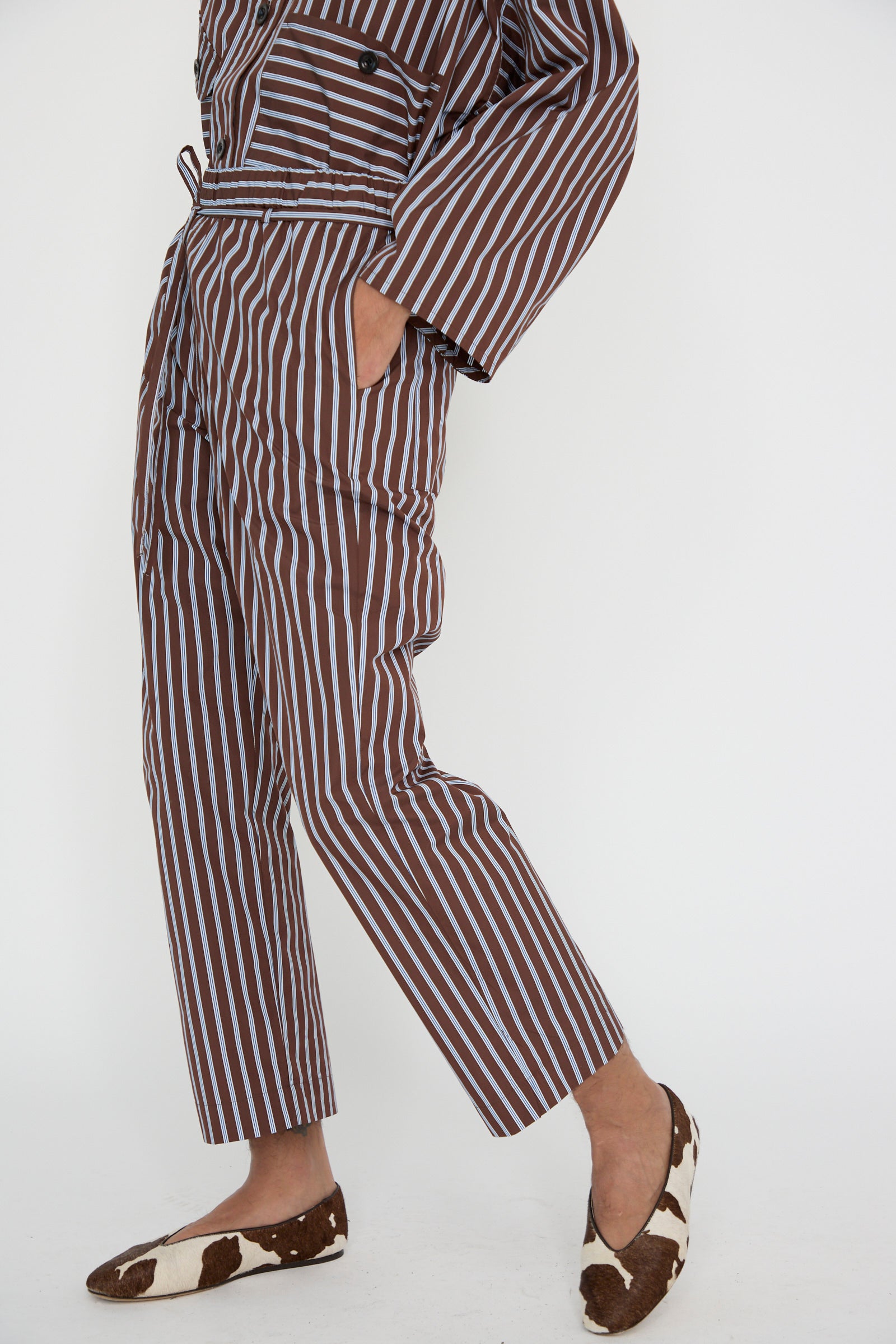 A person is wearing the KasMaria Taffeta Pajama Pants with Tie in Stripe, featuring a relaxed fit with an elasticated waist and  pockets, paired with cow-patterned slippers, show from torso down. 