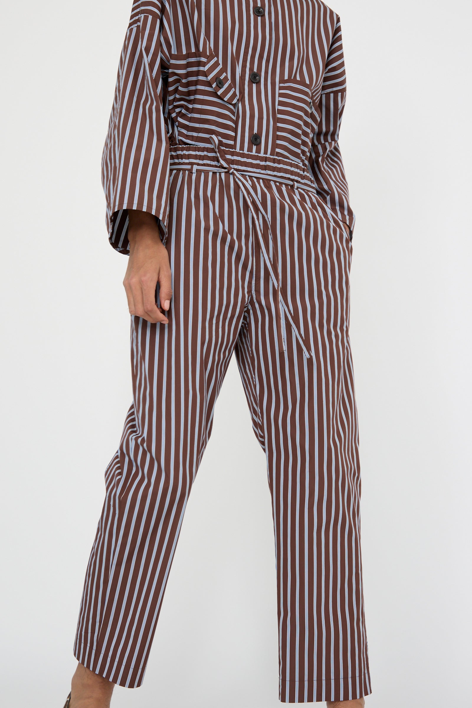 A person is seen against a plain background from below the neck, dressed in Taffeta Pajama Pants with Tie in Stripe from KasMaria, featuring a relaxed-fit design with matching brown and white stripes, made from Italian cotton taffeta and complemented by an elasticated waist.