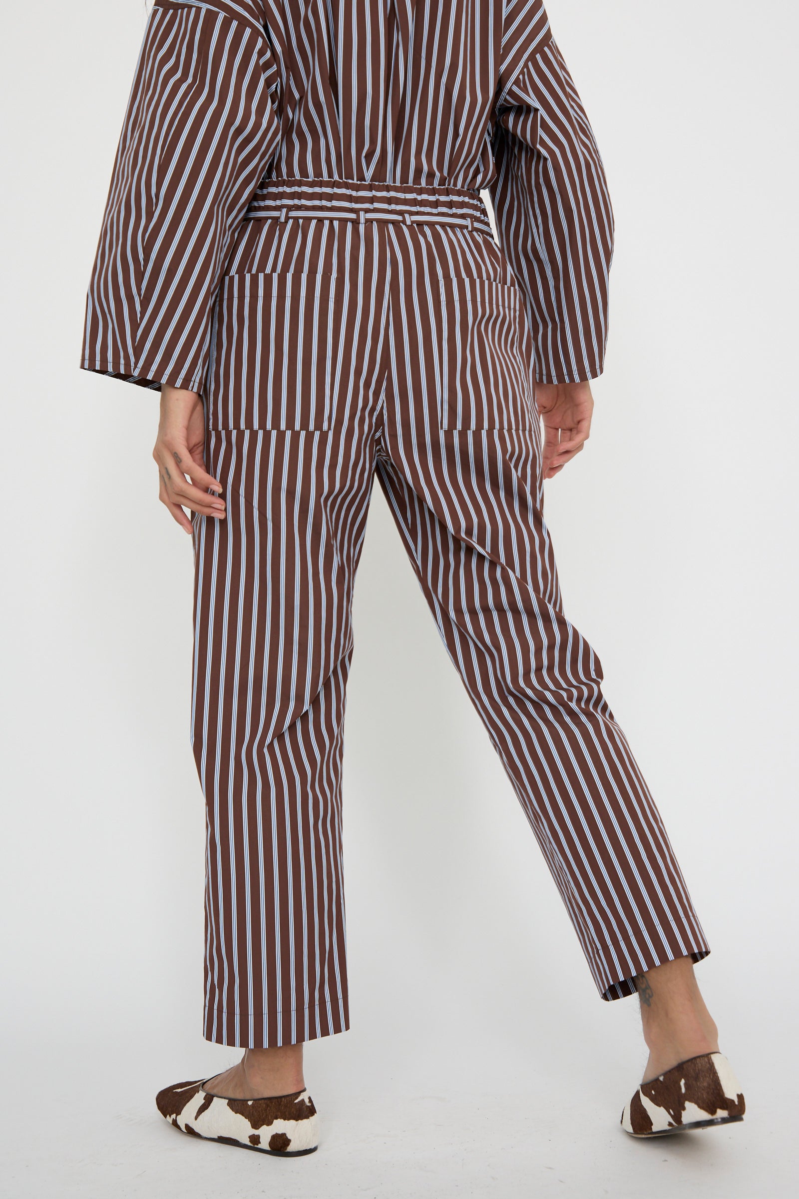 A person is seen from behind wearing the KasMaria Taffeta Pajama Pants with Tie in Stripe, crafted from luxurious Italian cotton taffeta featuring a relaxed elasticated waist in a brown and white stripe pattern, paired with cow-patterned shoes.