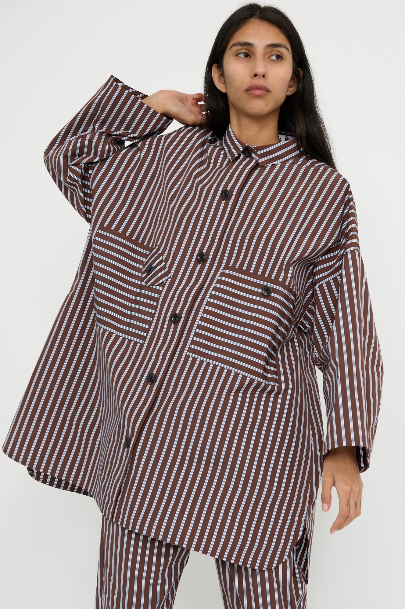 A person wearing the Taffeta Smock Shirt Dress in Stripe by KasMaria, featuring an oversized fit with brown and white stripes, stands against a plain background with one arm raised. 