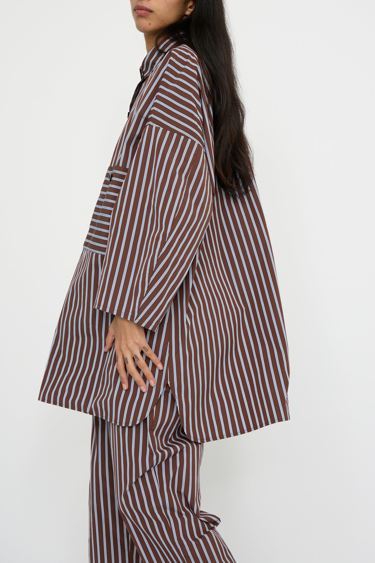 A person wearing the KasMaria Taffeta Smock Shirt Dress in Stripe, showcasing an oversized fit with brown and white Italian cotton taffeta, stands against a plain background and faces left. 