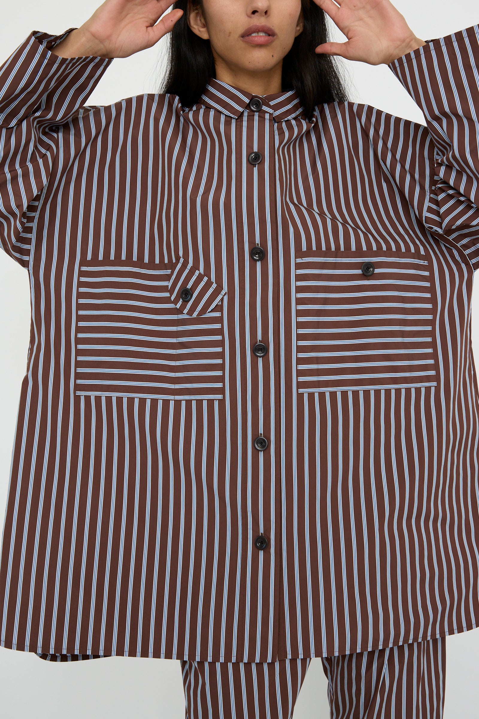 The individual wears a KasMaria Taffeta Smock Shirt Dress in Stripe, featuring an oversized fit with vertical and diagonal brown and white stripes, their arms raised.