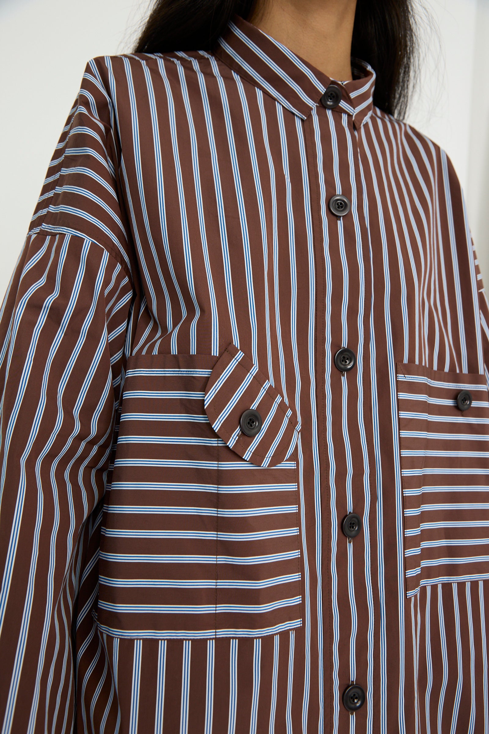 A close-up of an individual wearing the Taffeta Smock Shirt Dress in Stripe by KasMaria, highlights its oversized fit, crafted from Italian cotton taffeta with brown stripes, large pockets, and black buttons.