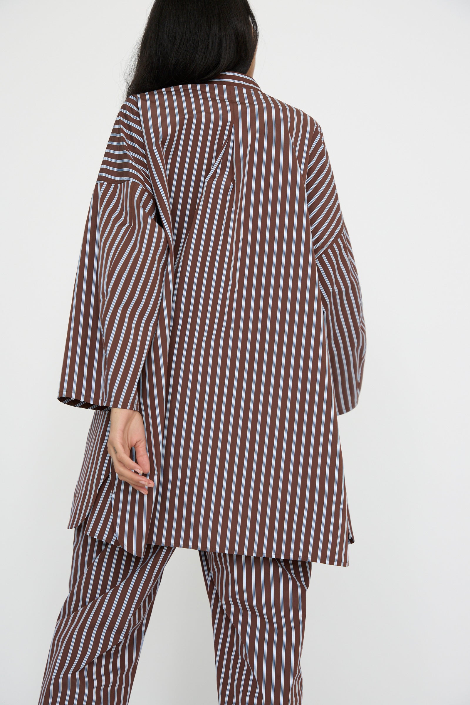 A person is seen in a KasMaria Taffeta Smock Shirt Dress in Stripe, featuring an oversized fit with brown and white stripes, facing away. 