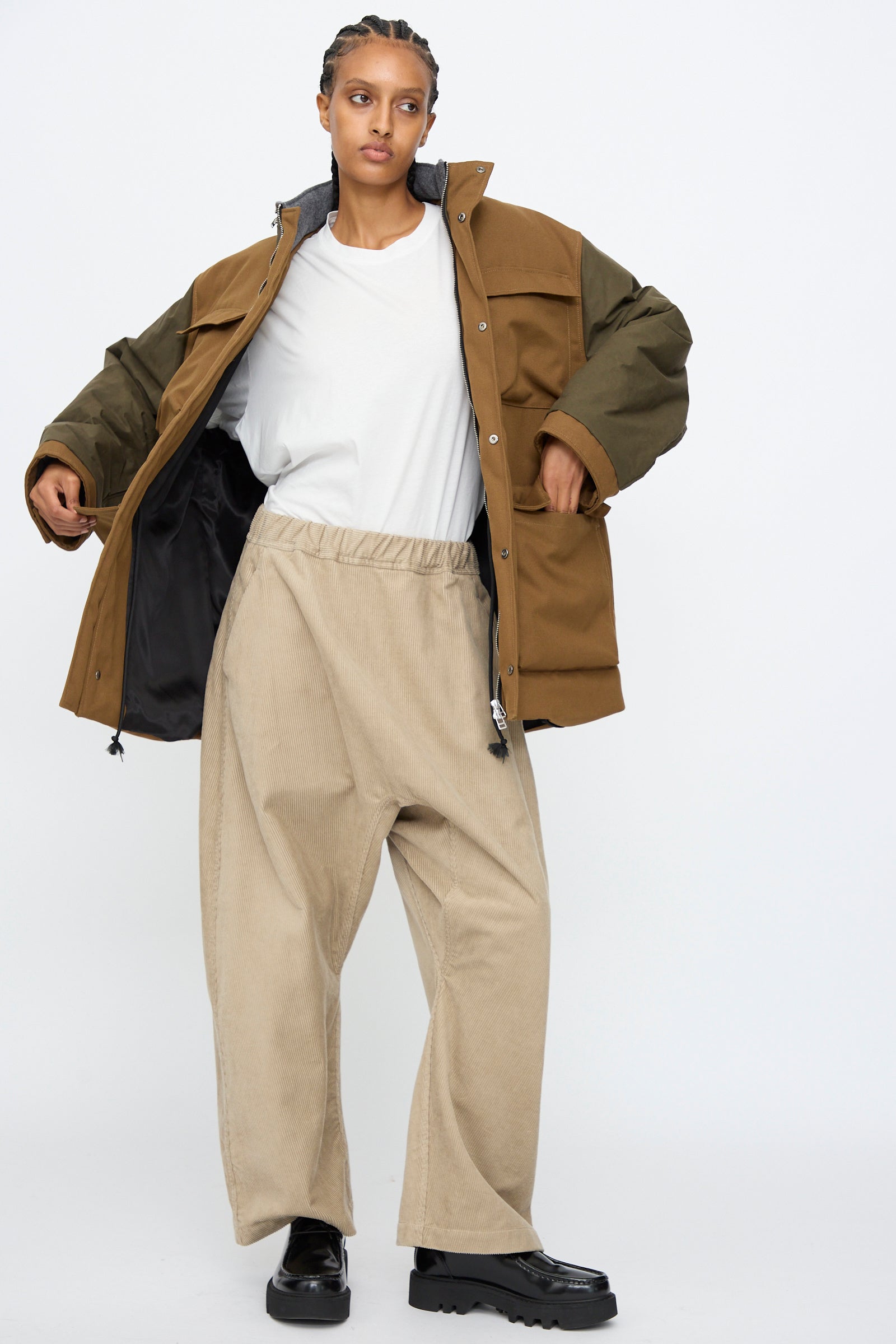 A person wears the Kassl Canvas Wax Field Jacket in Almond and Oil Black over a white shirt with beige wide-leg pants and black shoes, standing against a plain white background.