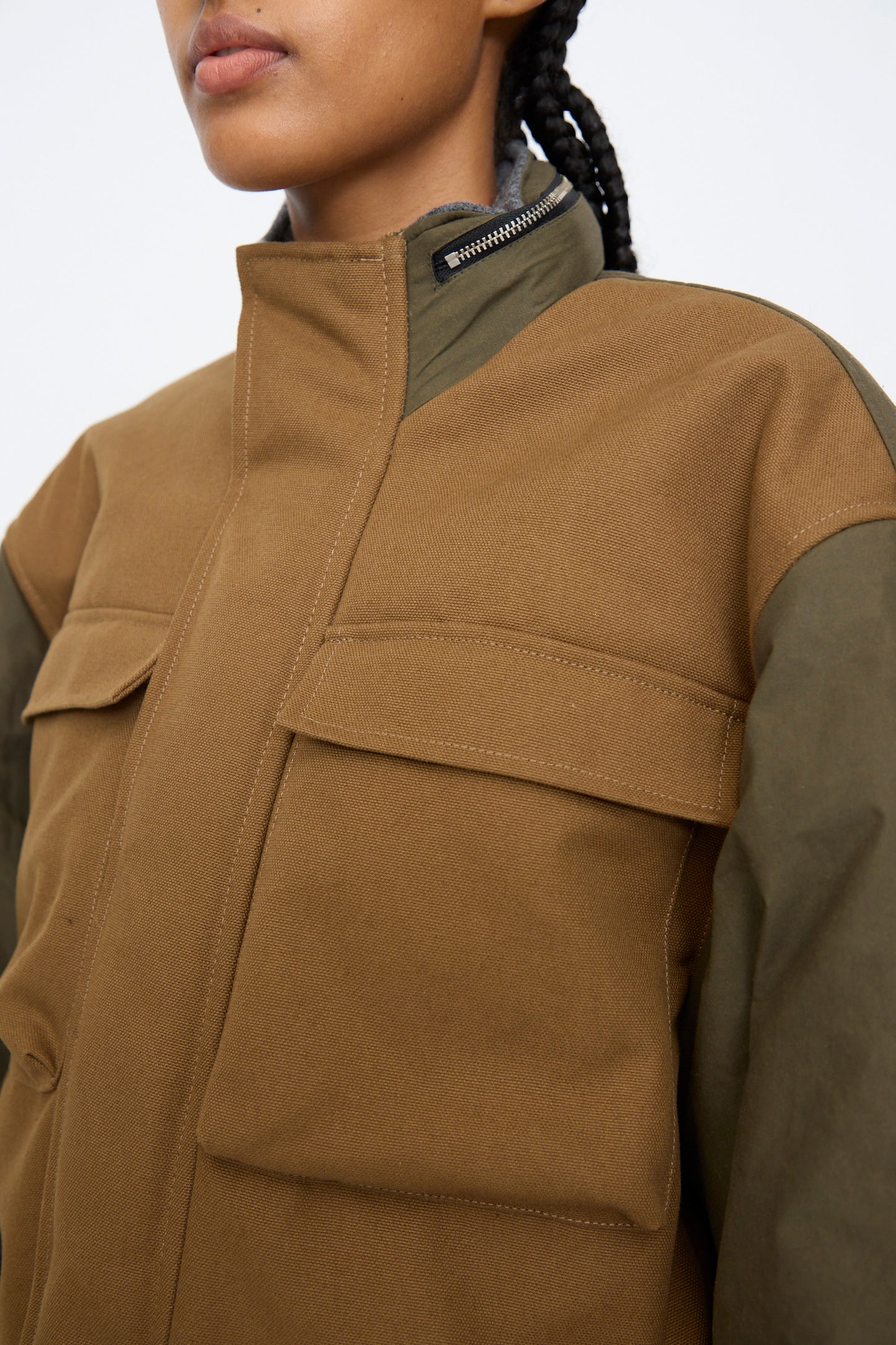 A person wears the Kassl Canvas Wax Field Jacket in Almond and Oil Black, featuring large front pockets and a high collar, shown from the shoulders up.